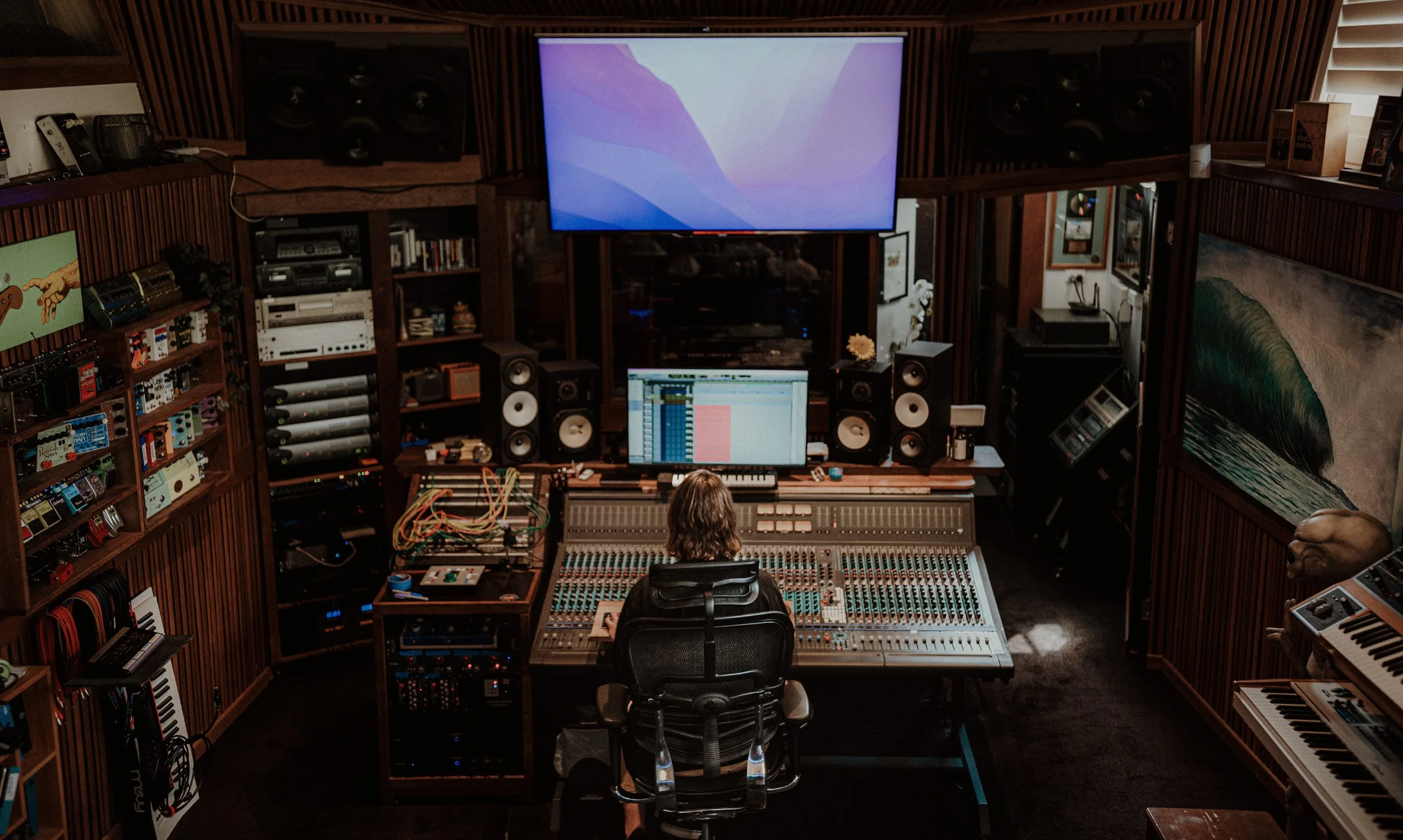 Inside a recording studio with a person at a mixing console, surrounded by speakers, musical instruments, and audio equipment.