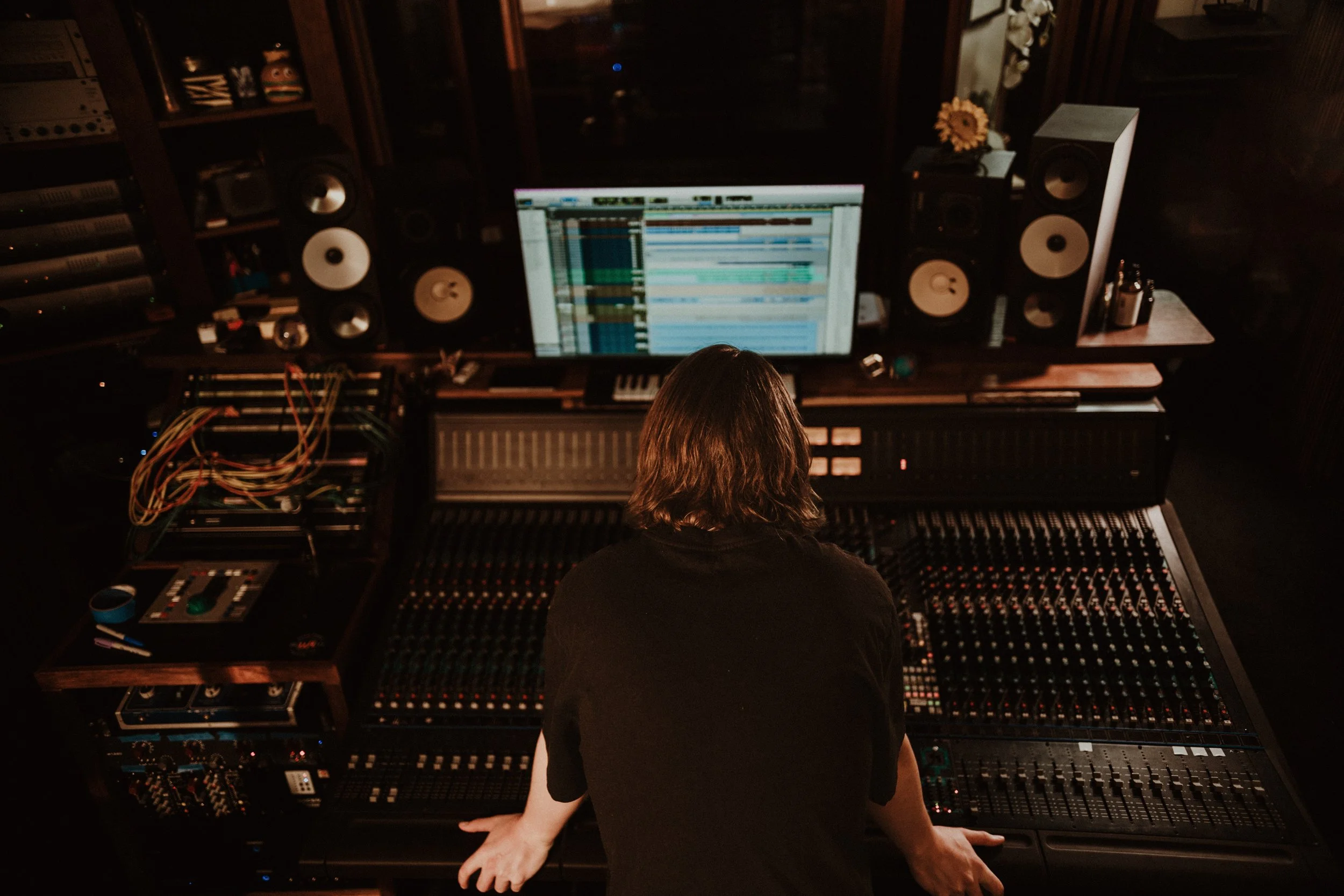 A person working at a large mixing console in a recording studio, with a computer monitor displaying audio tracks, surrounded by speakers and audio equipment.