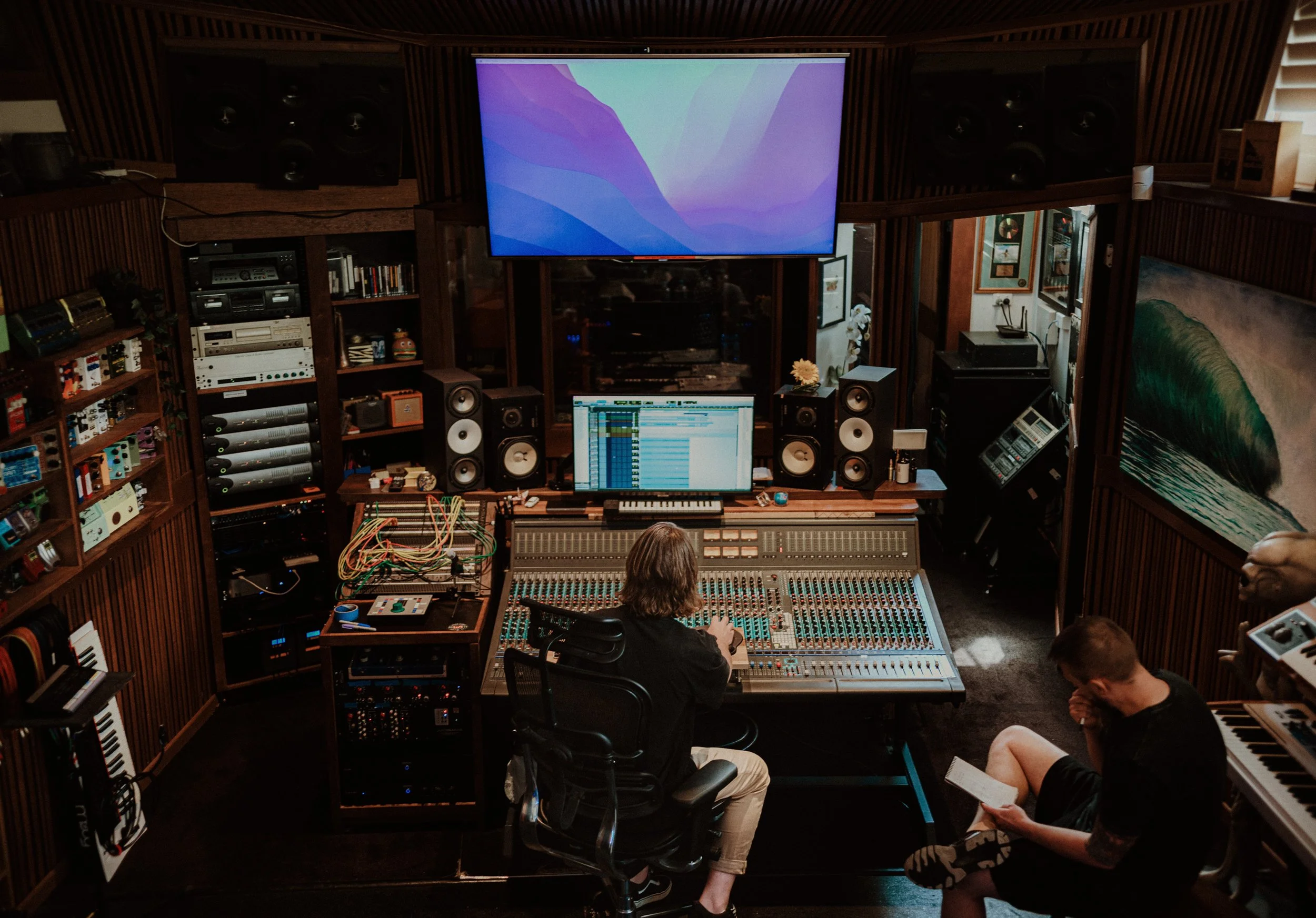 Recording studio with mixing console, computer screens, speakers, and two people, one at the mixing console and one reading a book.