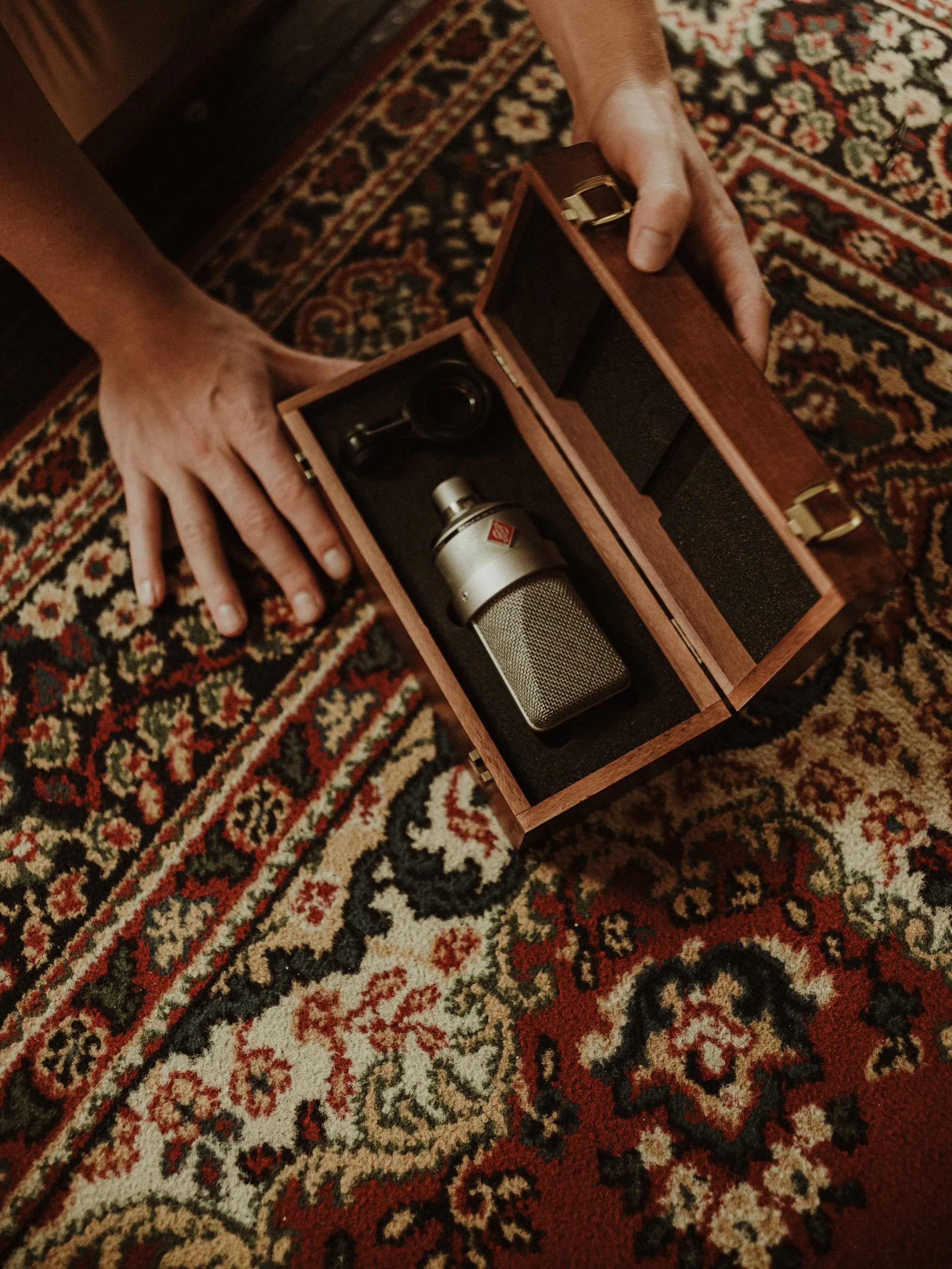 A person opening a wooden box containing a silver microphone and a small black item, on a patterned red and cream rug.