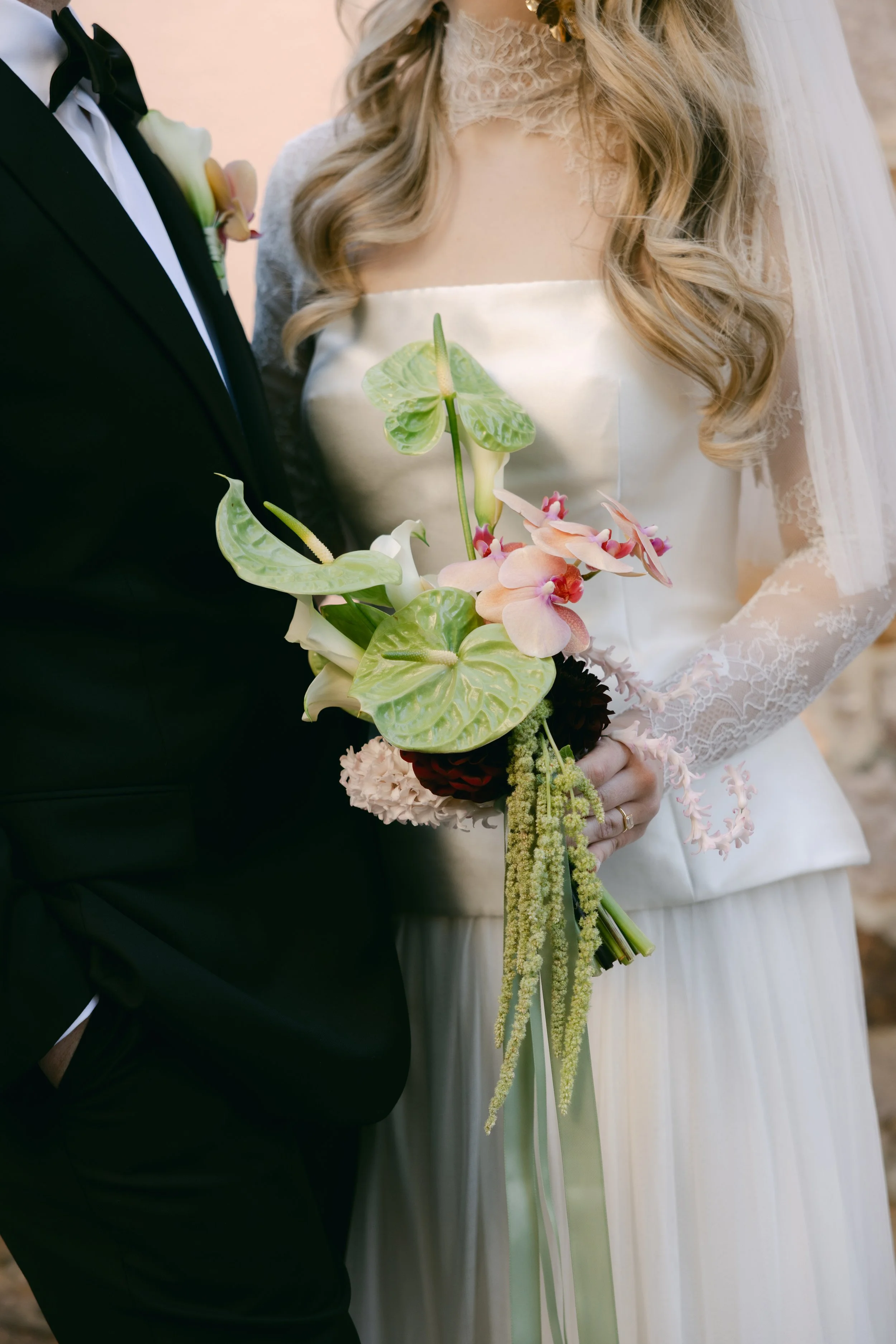 Close-up of bride and groom holding a floral bouquet at their wedding. The bride is wearing a lace wedding dress, and the groom is dressed in a black tuxedo with a bow tie.