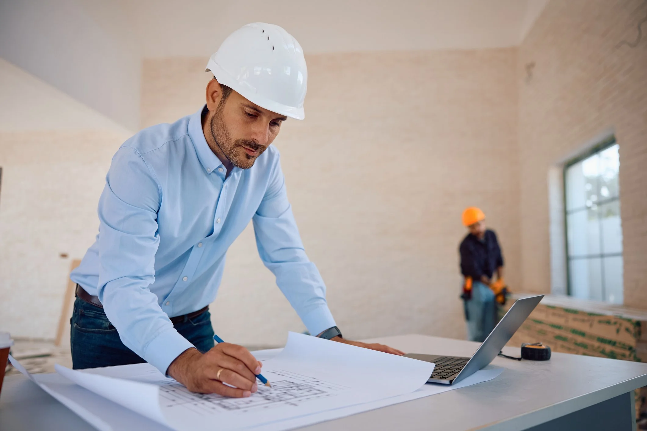 Person mit Schutzhelm prüft großformatige Baupläne auf einer Baustelle.
