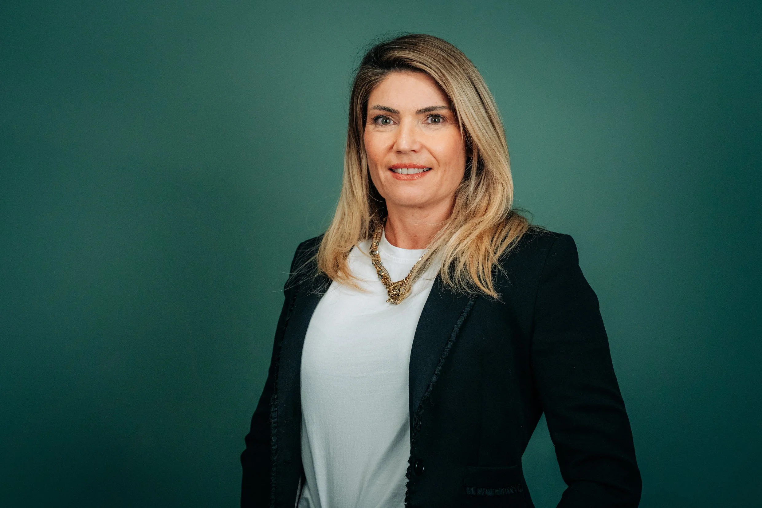 Porträt einer Frau mit blondem, schulterlangem Haar, schwarzer Blazer, weißem Top und goldener Kette vor grüner Hintergrund.