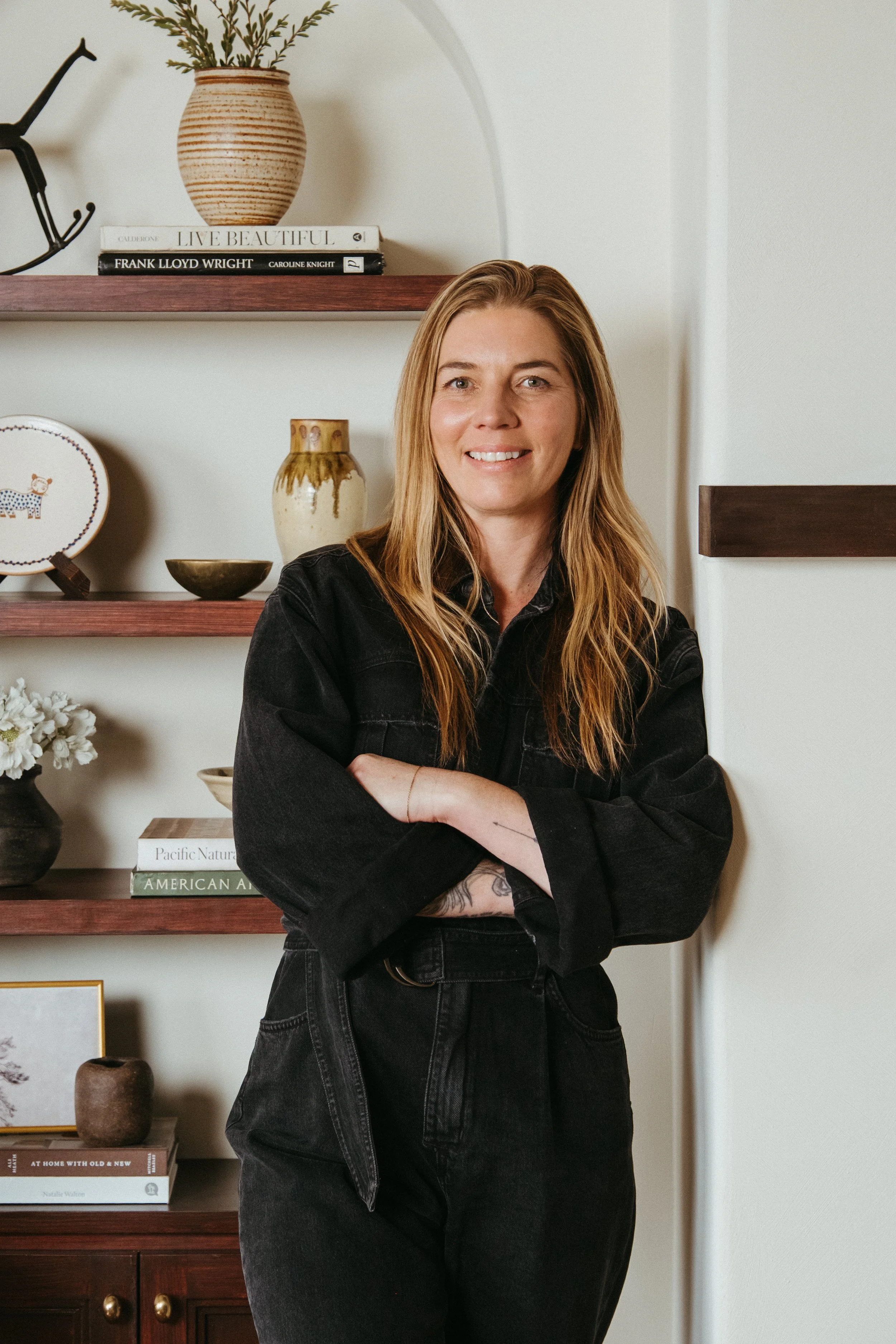 Person with arms crossed standing in front of a wooden shelf with books and decorative items.