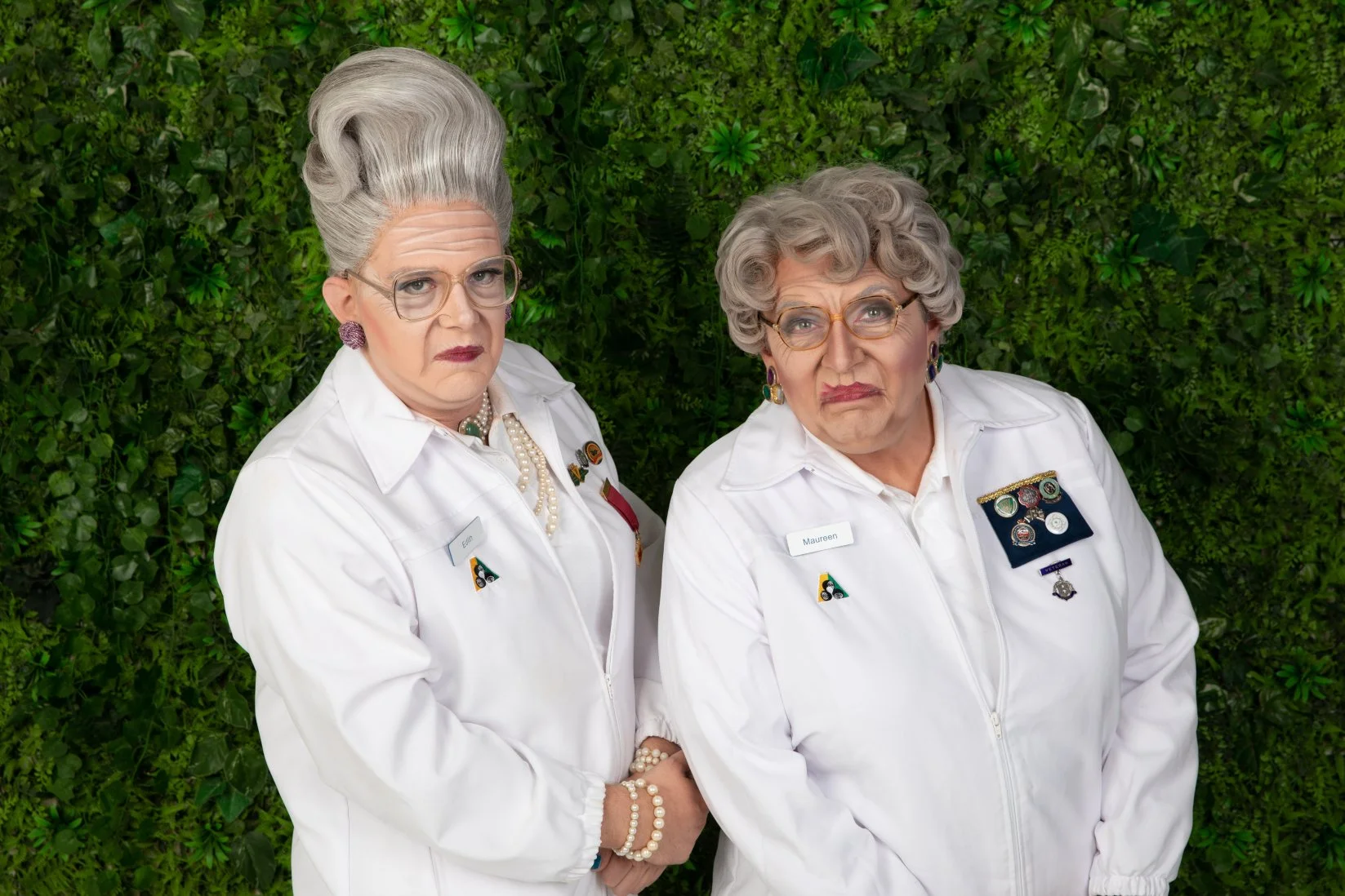 Two elderly women standing in front of a green leafy background, wearing white uniforms with badges and medals, making serious and unamused expressions.
