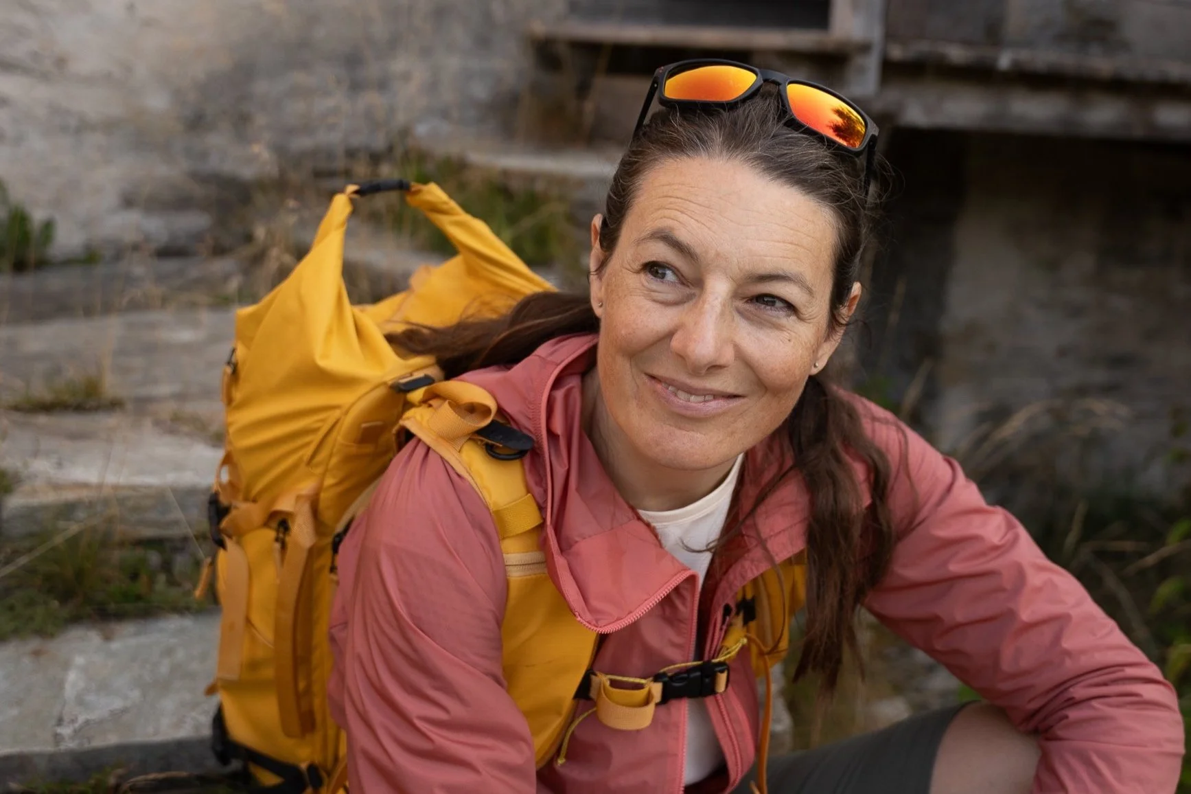 Smiling woman with long hair wearing sunglasses on her head, a pink jacket, and a yellow backpack sitting outdoors.