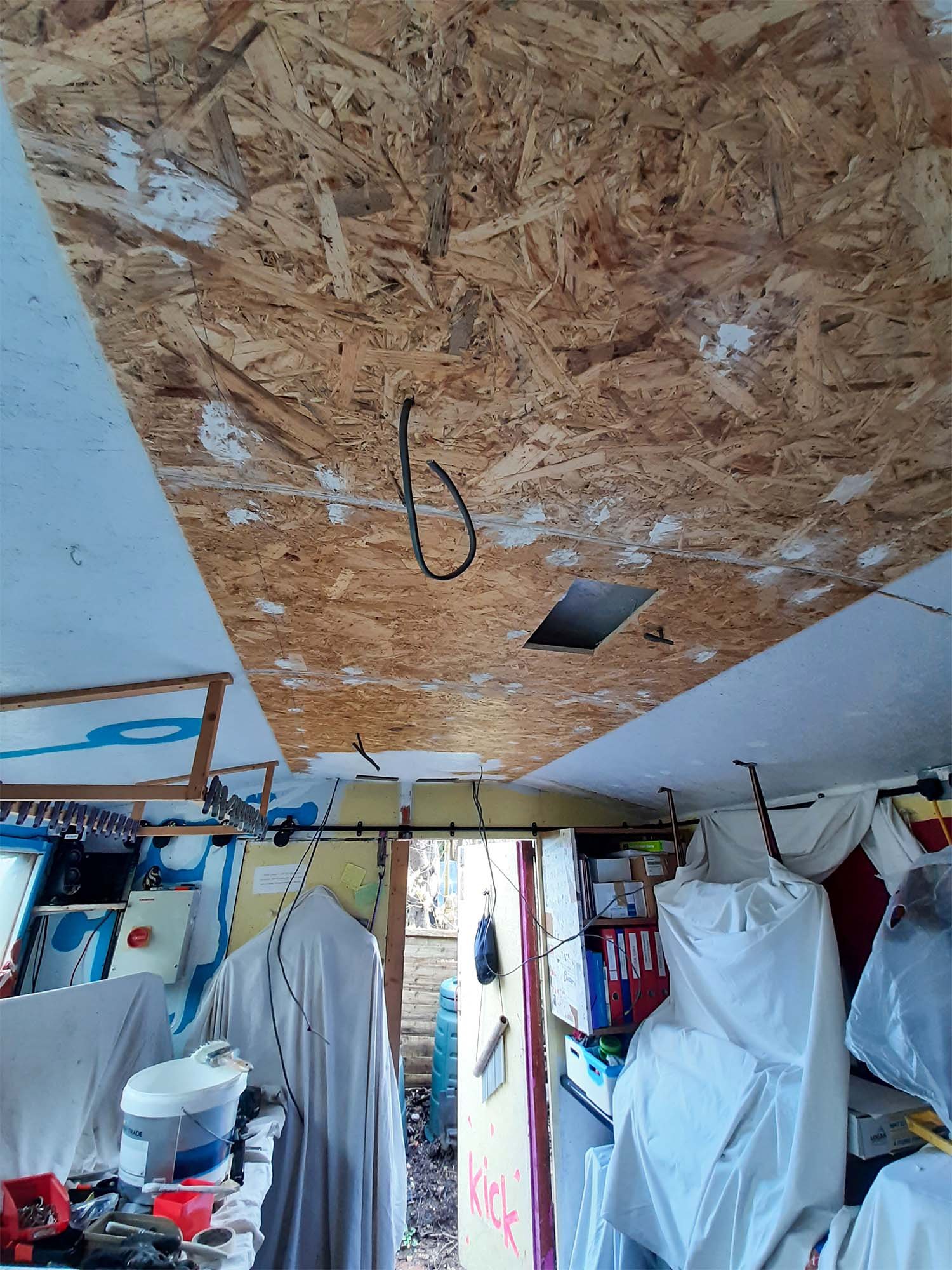 interior of studio with wooden sheet material applied to ceiling mid-renovation. Cables poke through holes in the material and signs of filler are available. White sheets cover a majority of visible surfaces