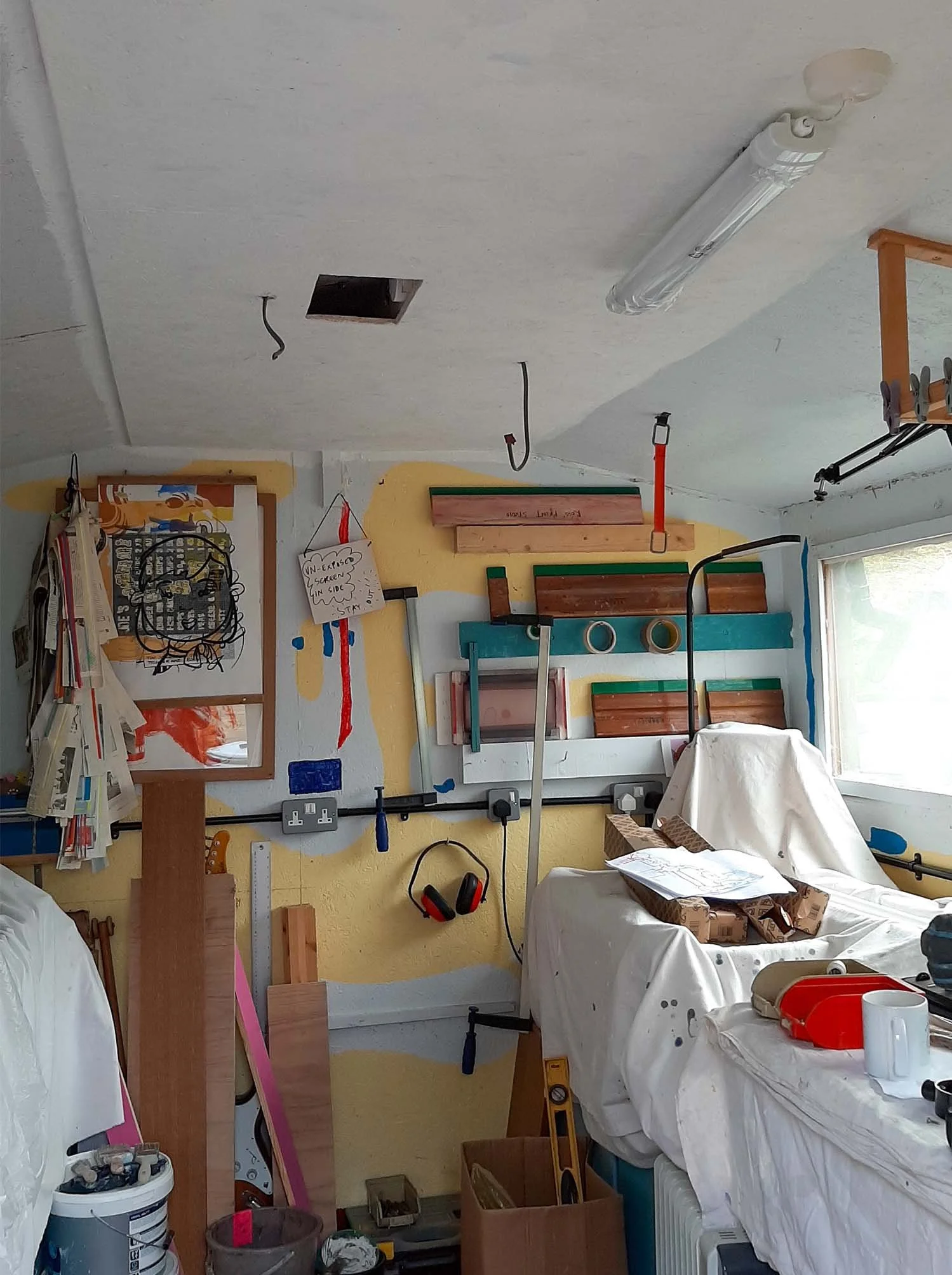 studio interior with white sheets covering various surfaces and DIY materials scattered about. Cables poke through holes in the newly-painted ceiling on which new likely fixtures have been affixed.