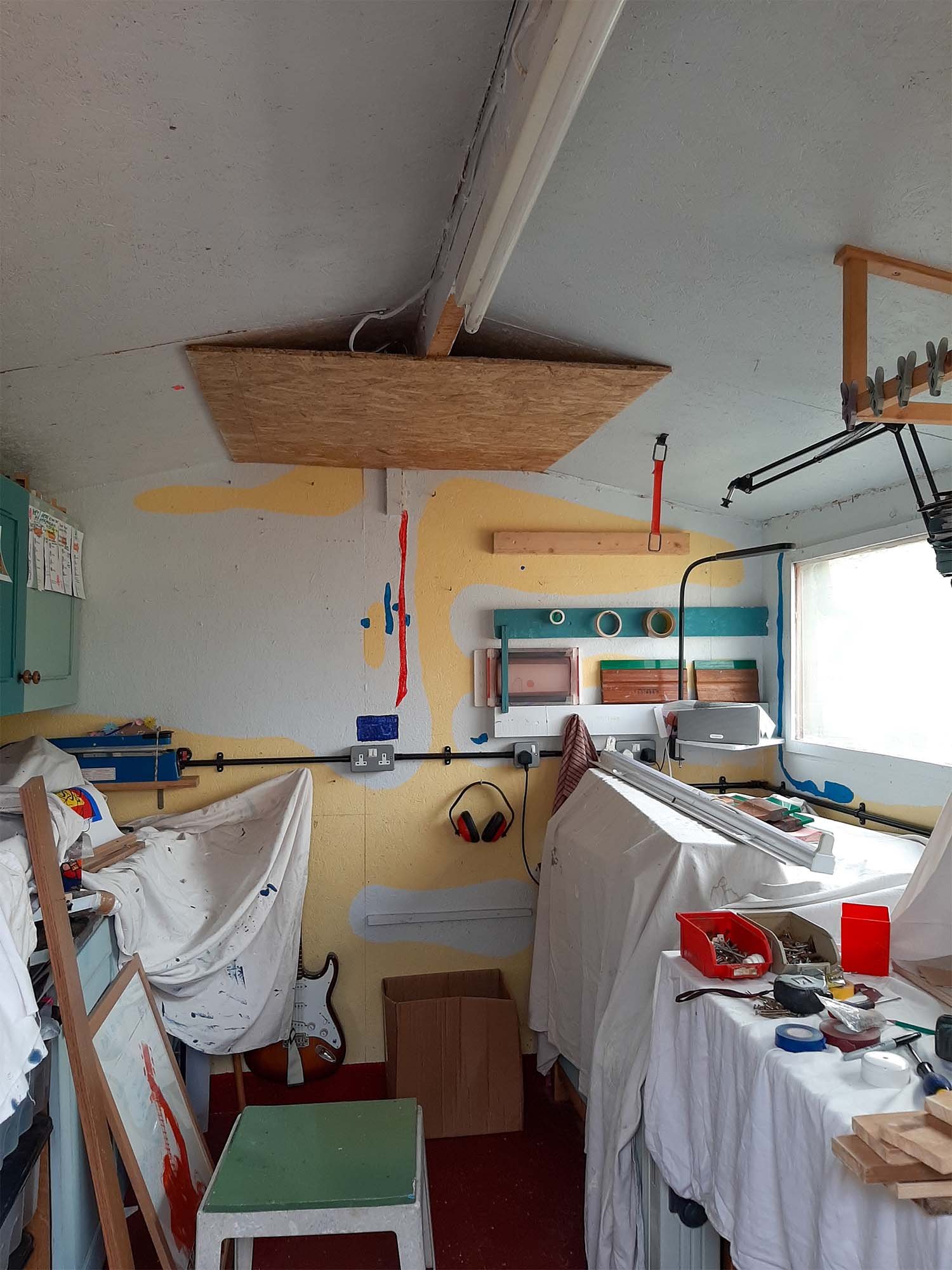 interior of studio with wooden sheet material applied to ceiling mid-renovation. Sheets covering work surfaces on top of which tools and materials are strewn