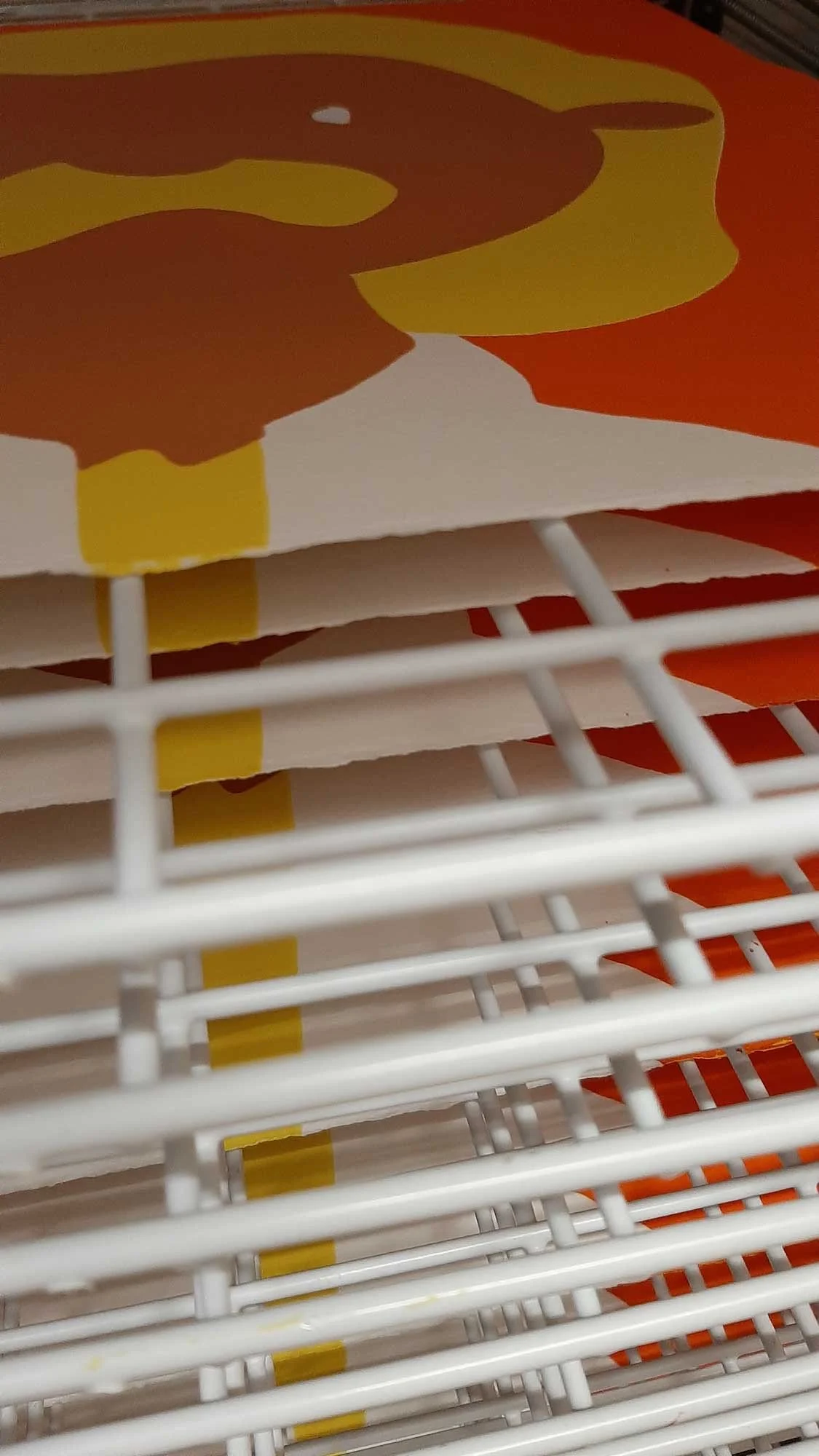 Multiple silkscreen prints drying on a white metal rack, featuring bold abstract artwork in orange, yellow, and brown.
