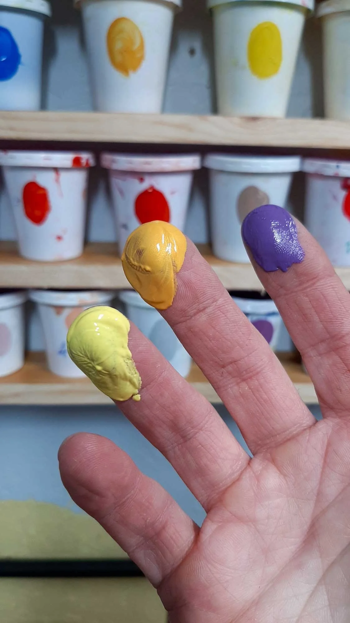 Close-up of a hand with yellow, orange, and purple paint on fingertips, in front of shelves of color-coded mixing cups.