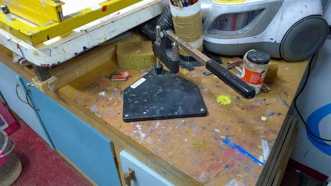 A cluttered workbench with a button press, screen frame and tools used for framing and pinning prints.