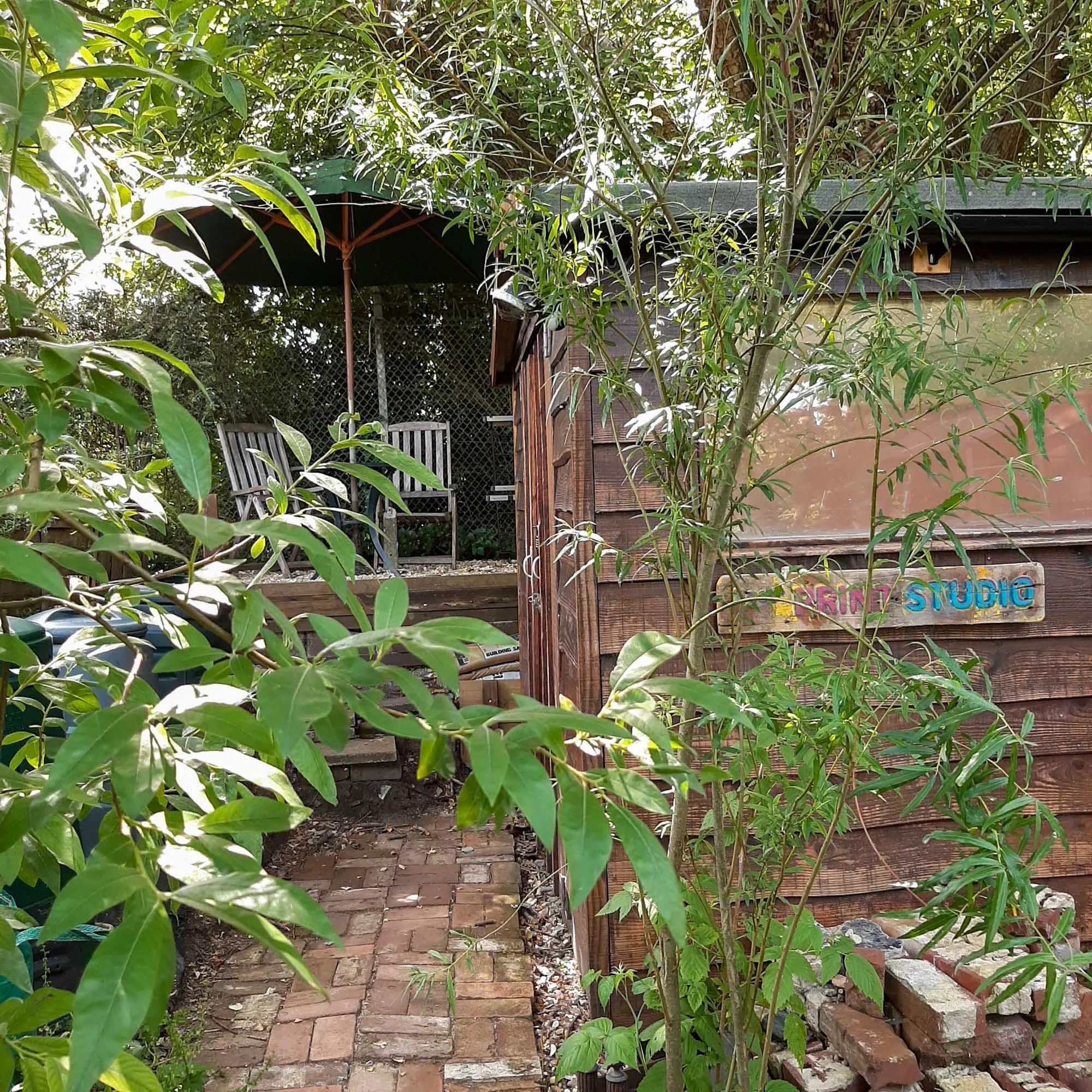 Wooden garden shed with a colorful “PRINT STUDIO” sign, surrounded by greenery and a brick path leading to the entrance.