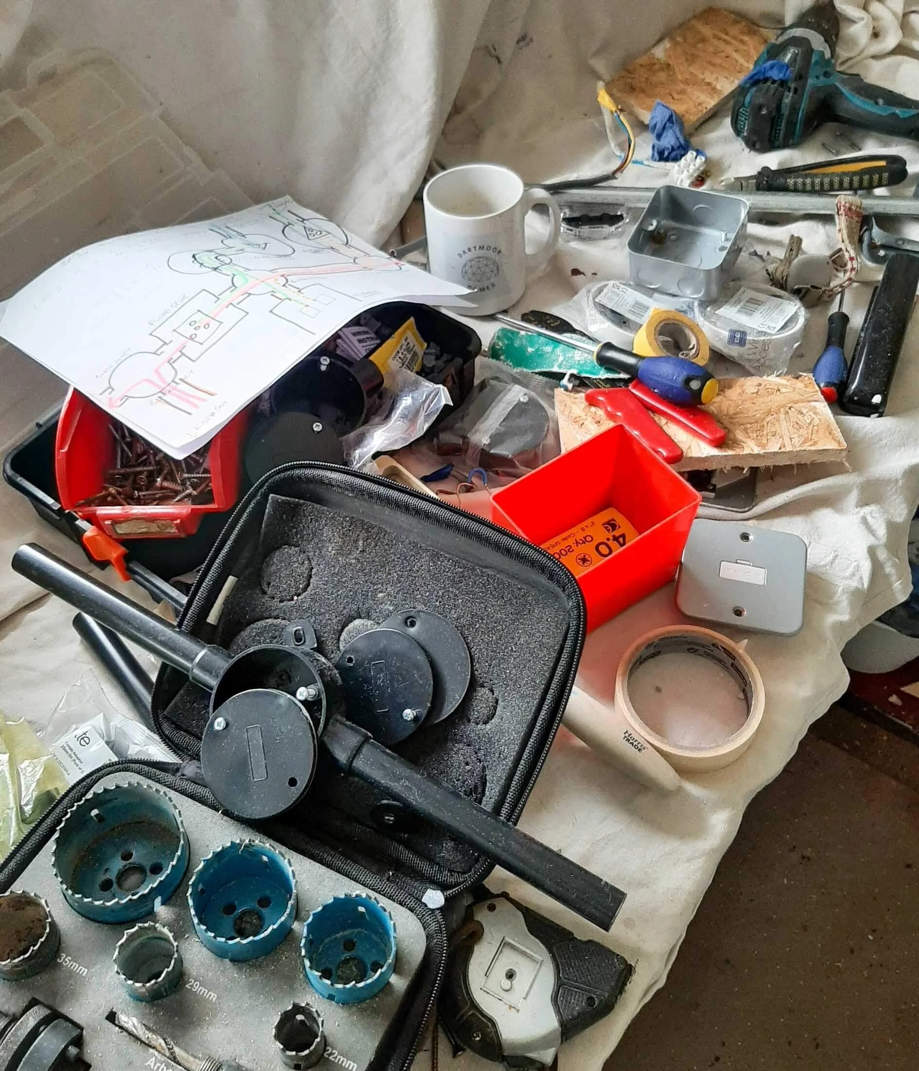 A table covered with tools, drill bits, sandpaper and a wiring diagram used during studio electrical planning.