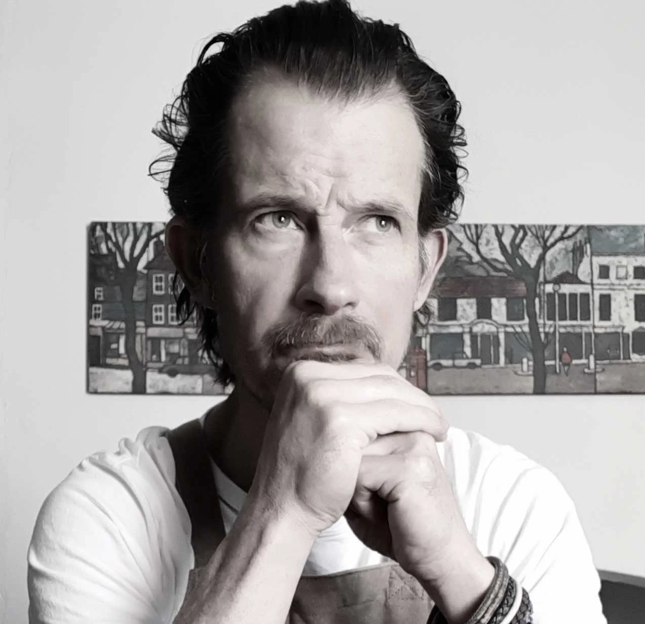 A man with dark wavy hair and a goatee, resting his chin on his fists, with a thoughtful expression, in front of a painting of a street scene with trees and buildings.