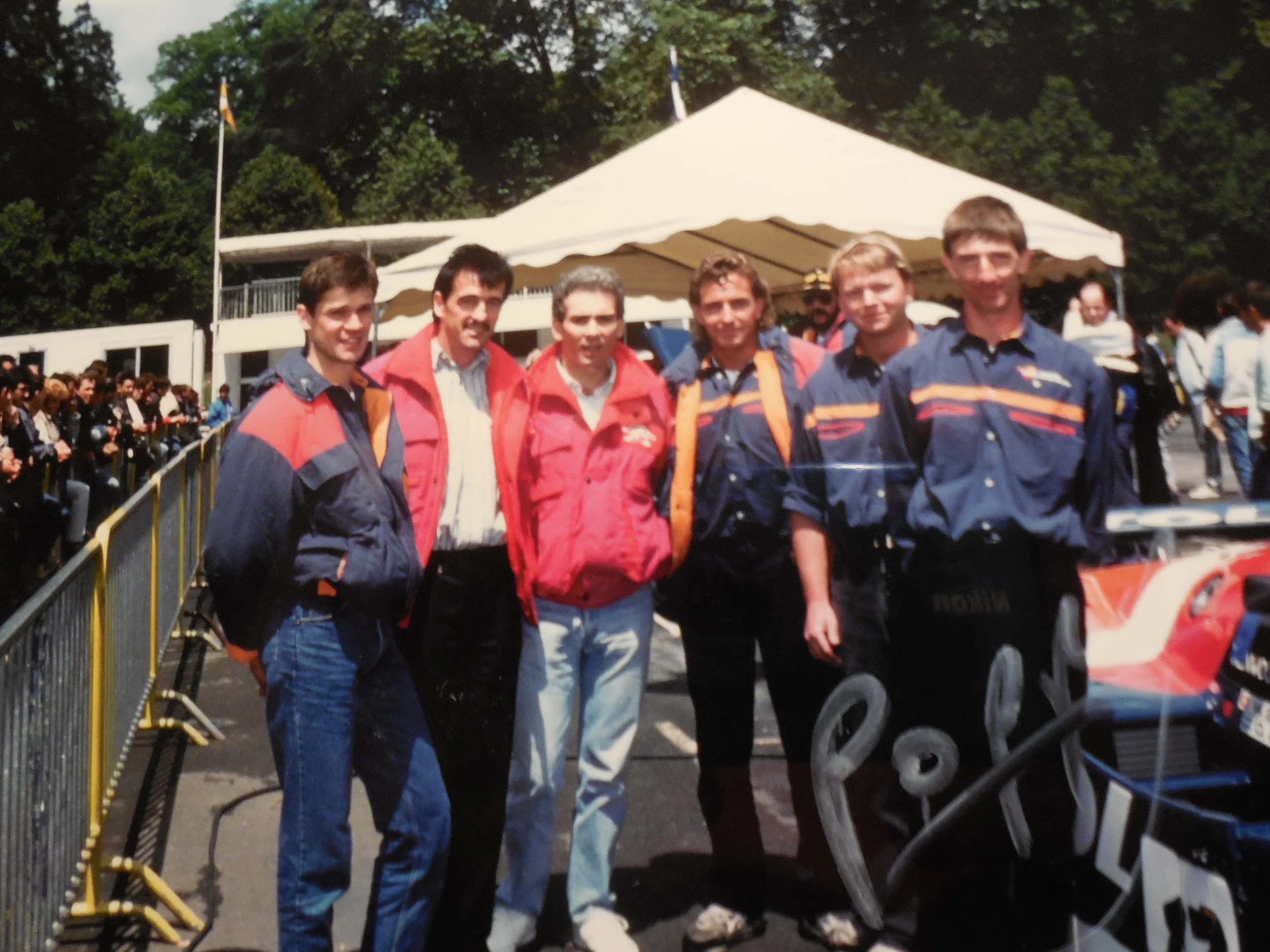 Fünf Menschen stehen vor einem weißen Zelt bei einem Motorsport-Event, die Mehrheit trägt kolorierte Rennteamjacken.