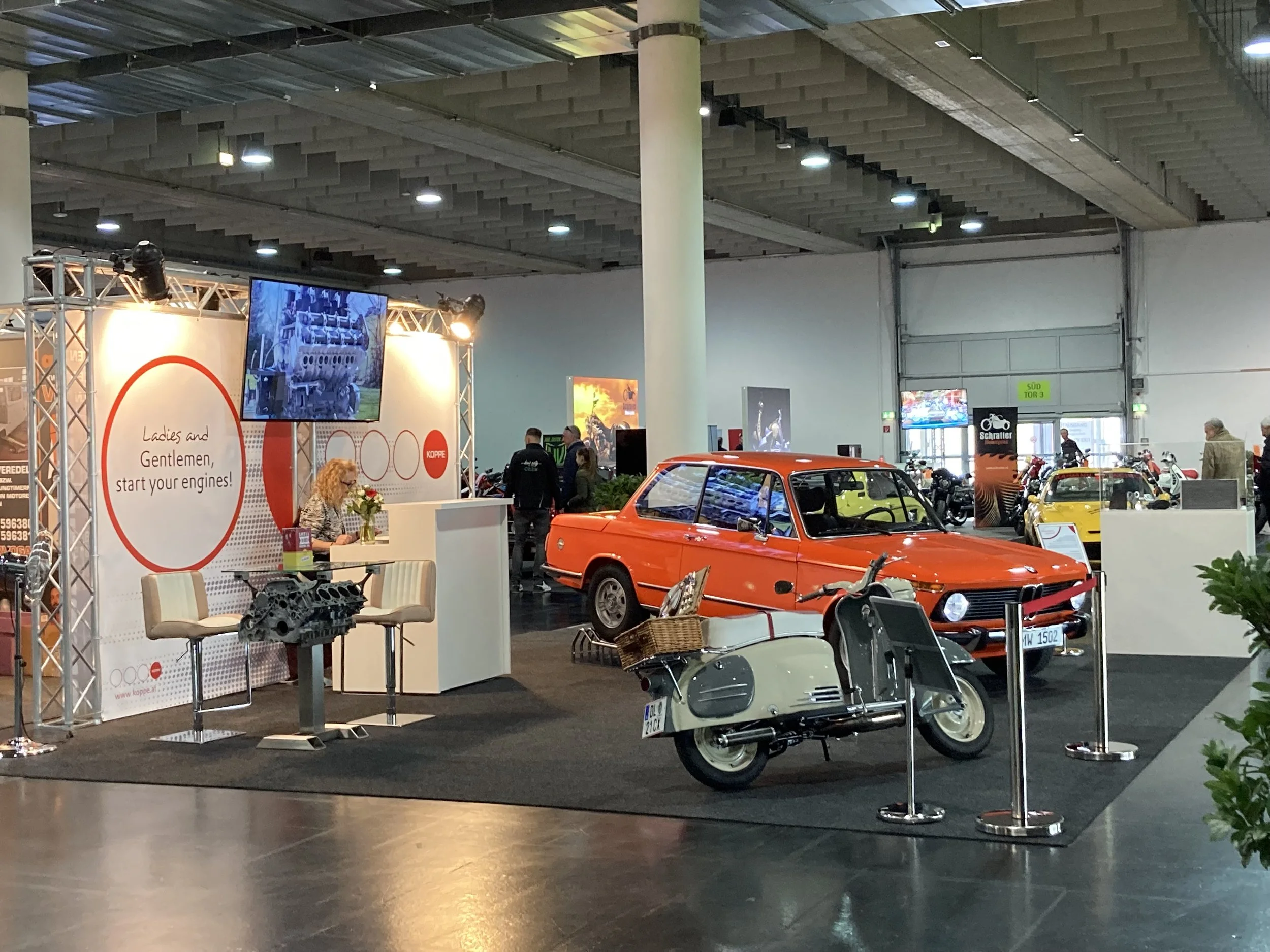 Auto- und Motorradmuseum mit einem orangefarbenen klassischen Auto und einem weißen Roller, Ausstellungsstand mit einem Bildschirm und einem Empfangstresen, im Hintergrund weitere Fahrzeuge und Aussteller.