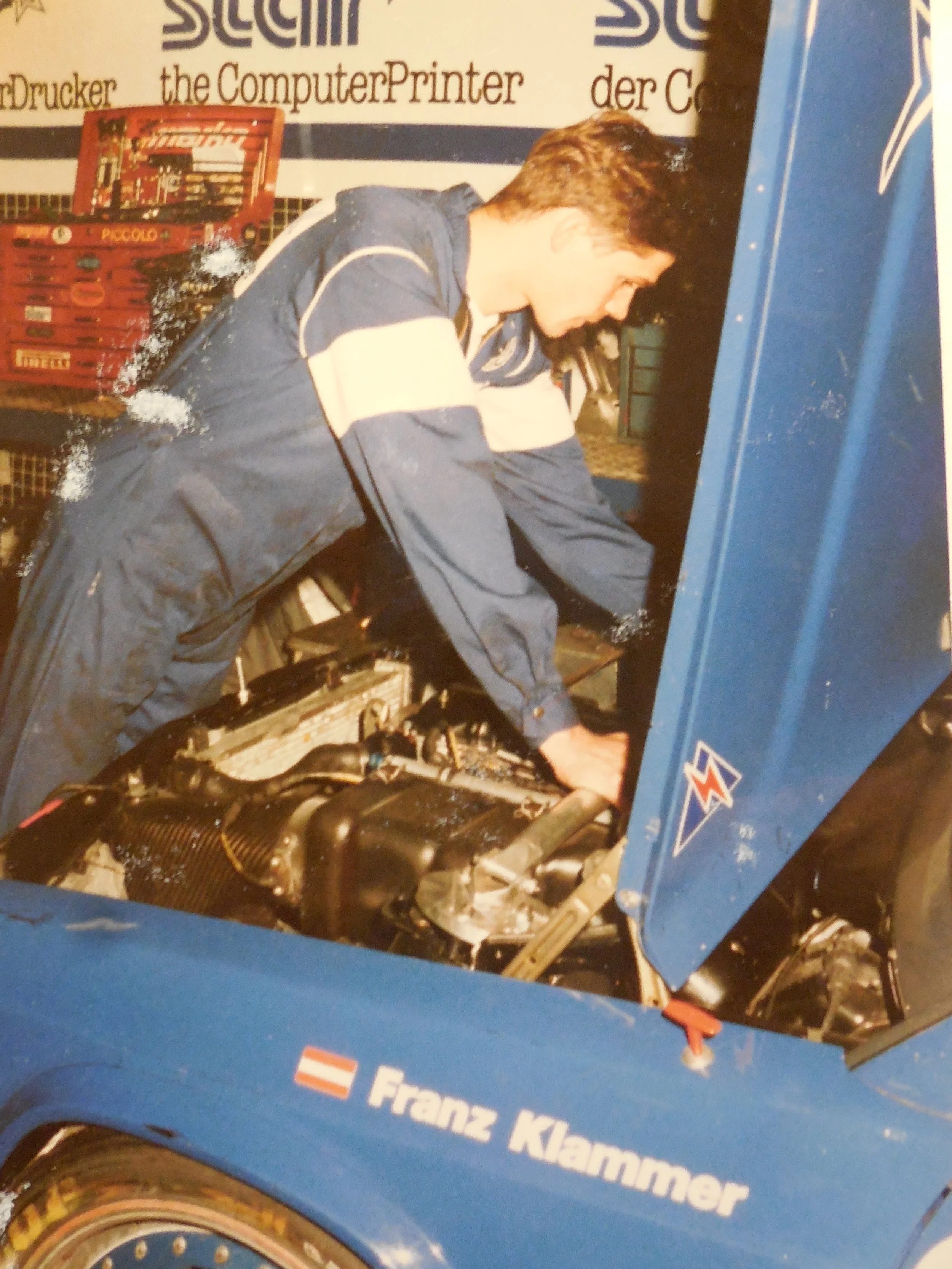 Ein junger Mann in blauer und weißer Rennfahrerjacke arbeitet an einem blauen Rennauto. Der Wagen hat den Namen 'Franz Klammer' auf der Seite. Die Szene scheint in einer Garage oder Werkstatt stattgefunden zu haben.