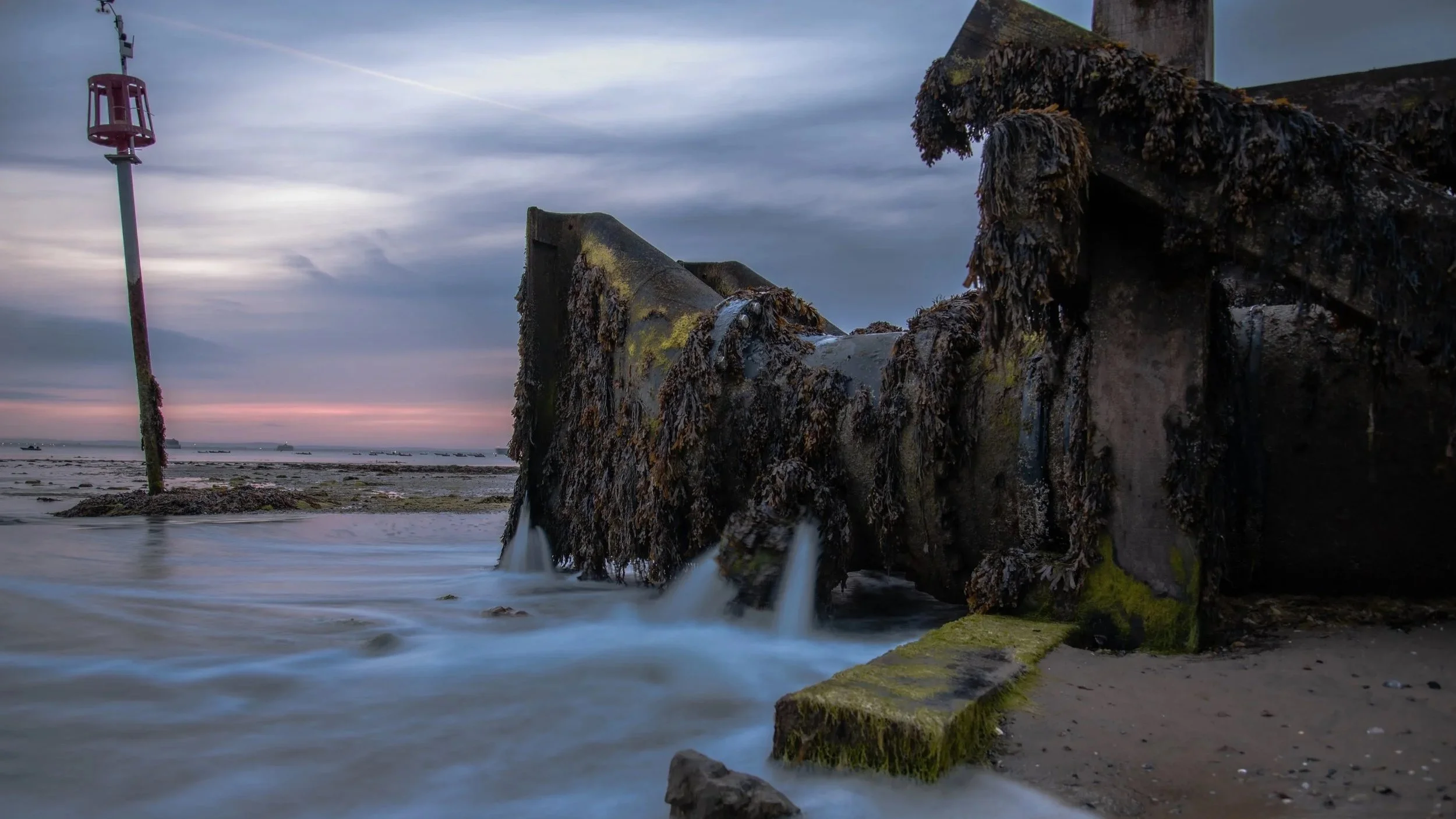 Old, moss-covered concrete structure on a beach with water flowing over it, a pink lighthouse on a pole, and a cloudy sky at sunset.