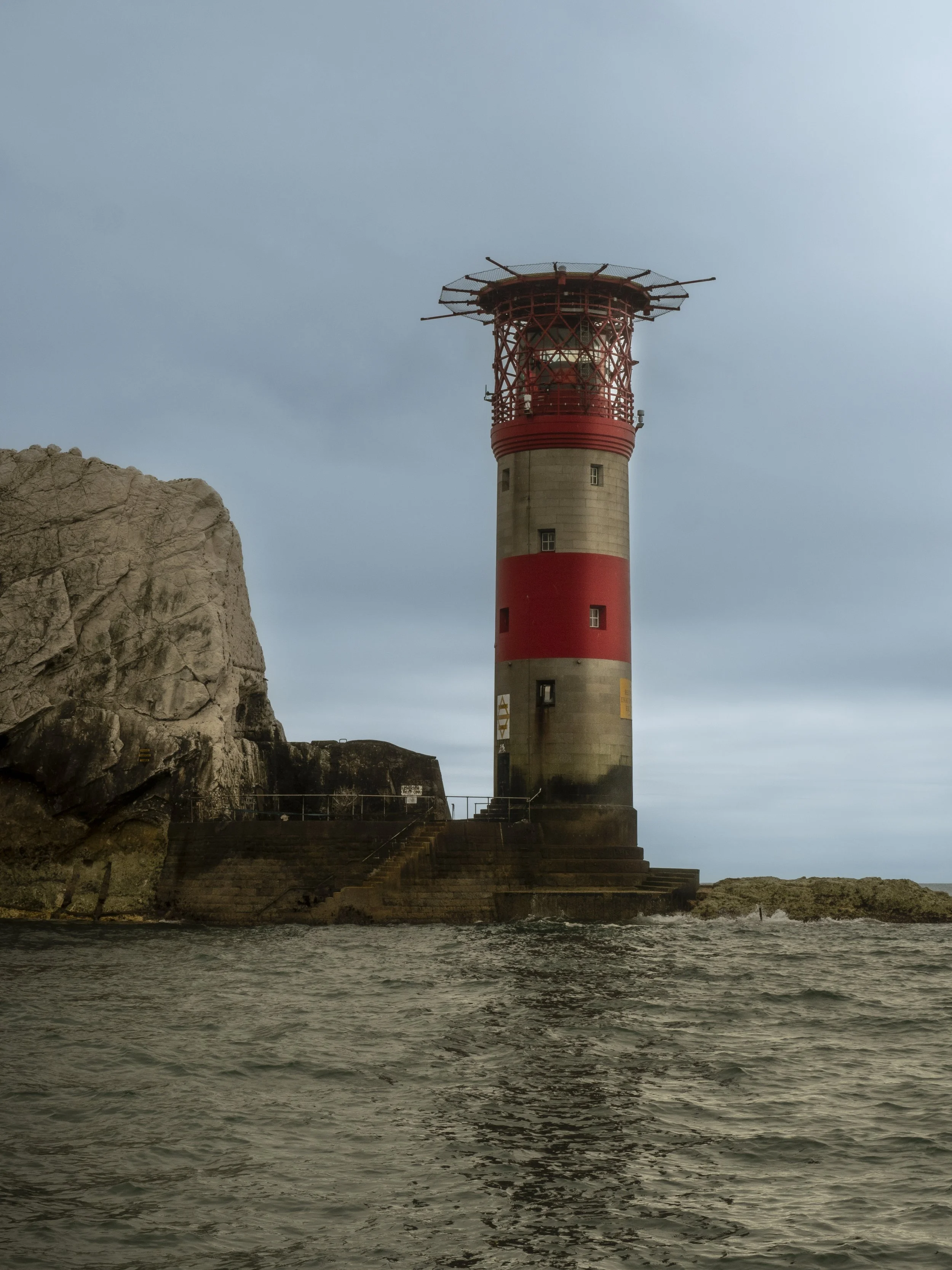 A lighthouse on a rocky shoreline with steps leading up to the entrance, set against a cloudy sky.