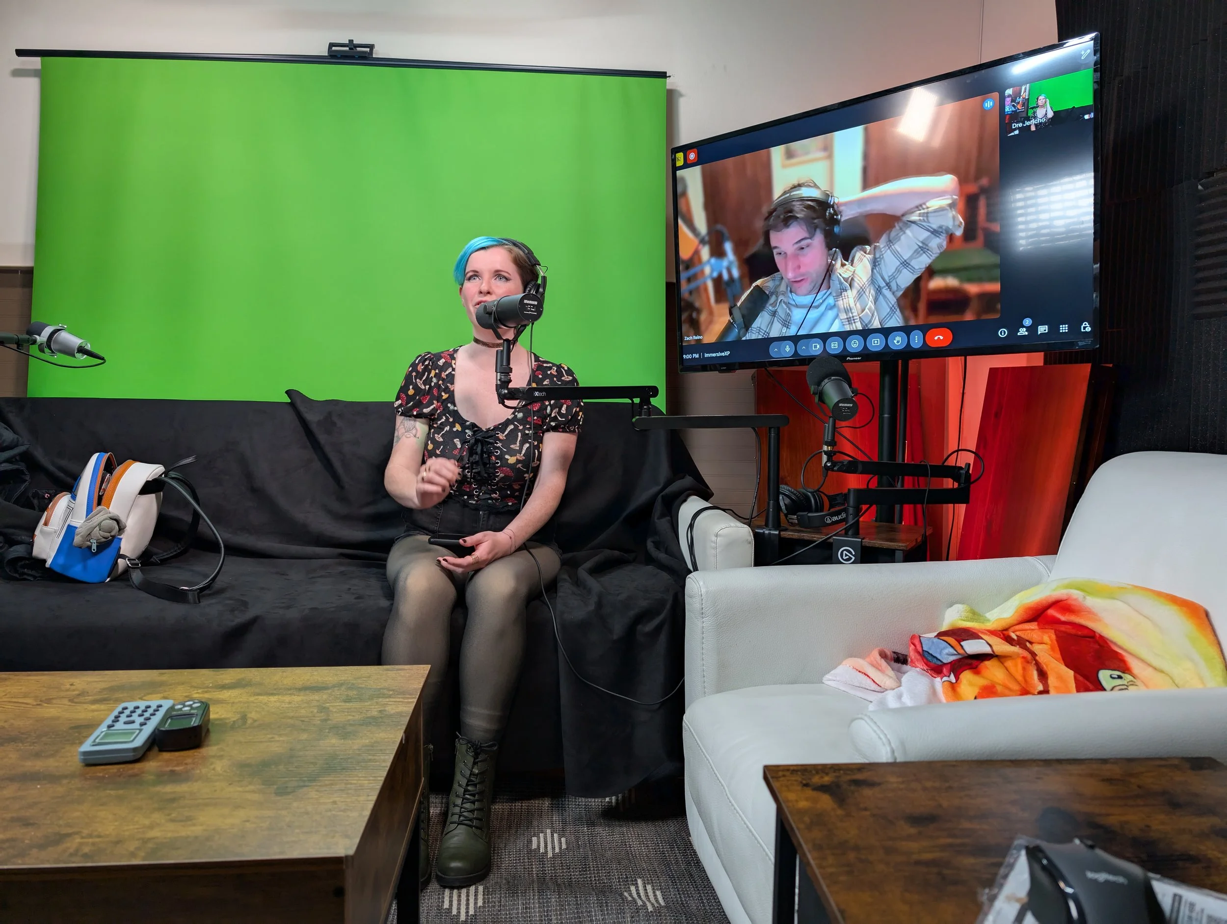 A woman with short, blue hair and tattoos on her arm sits on a black-covered couch, speaking into a microphone, while a man with headphones appears on a large screen in a video call. The room has a green screen behind her and a wooden coffee table in front of her.