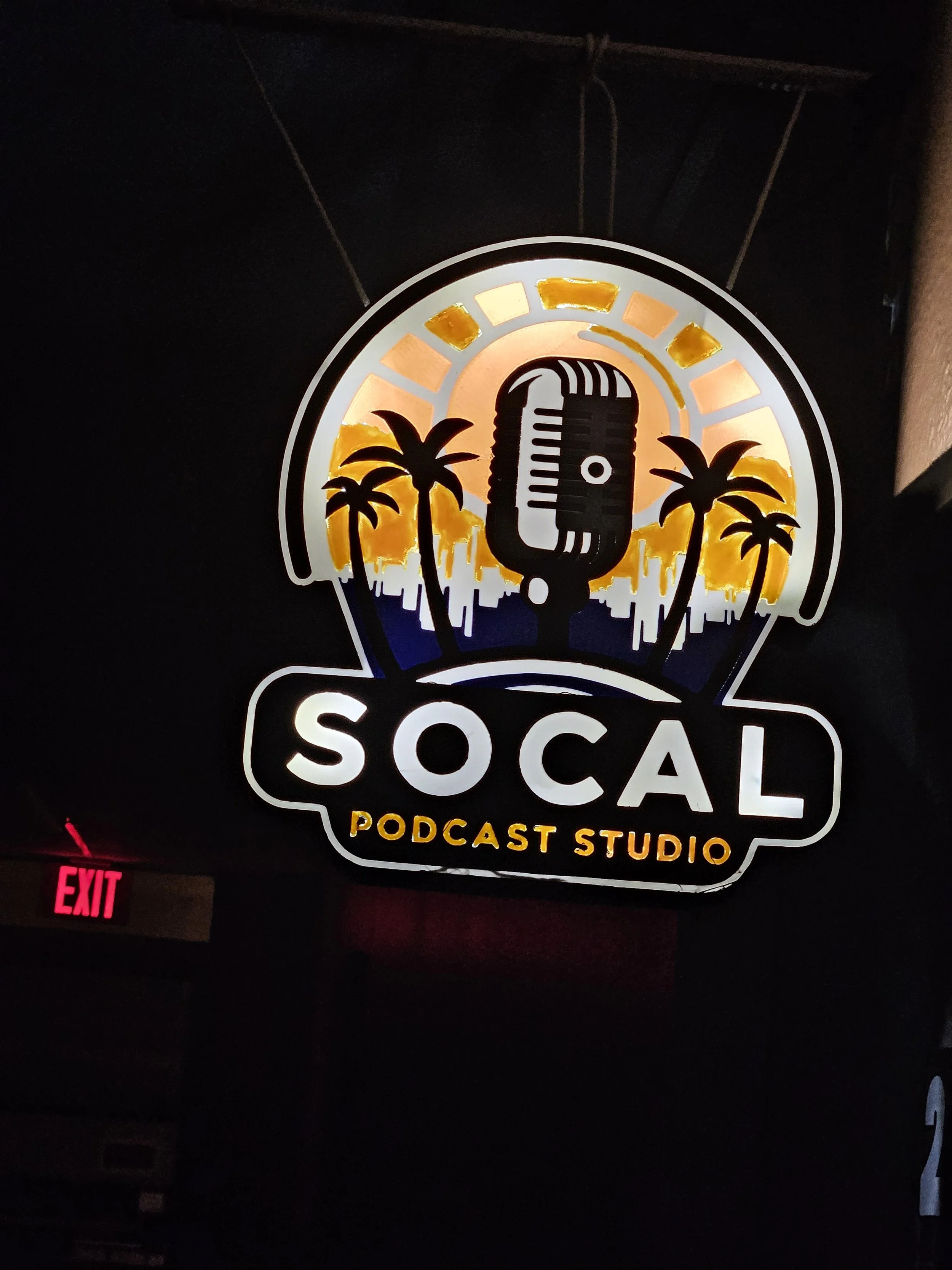 Neon sign for SOCAL Podcast Studio featuring a vintage microphone, palm trees, and a cityscape silhouette with a sunset background.