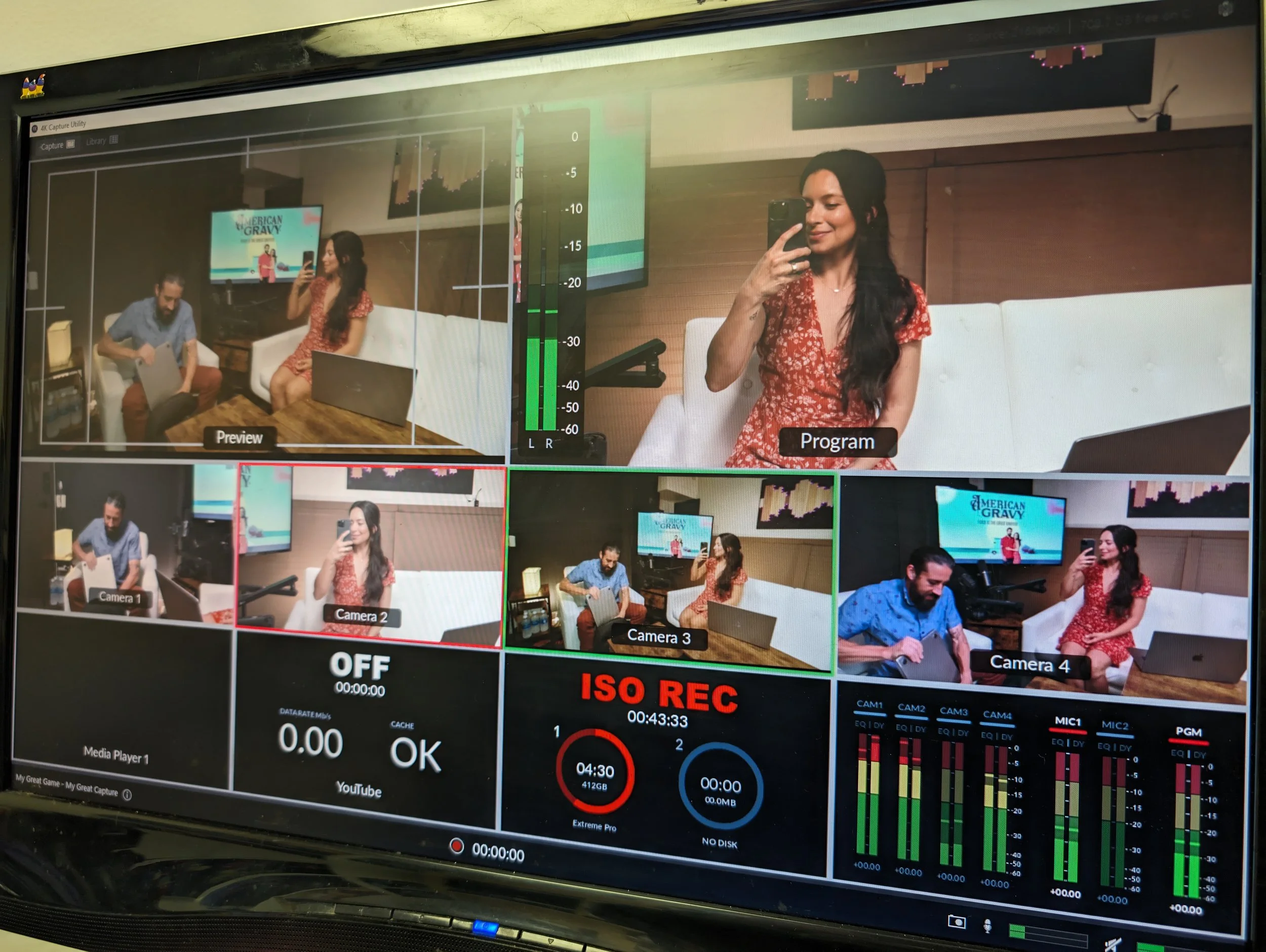 A computer monitor displaying a live video feed of a woman in a red floral dress taking a selfie, with multiple camera views, audio levels, and recording information on the screen.