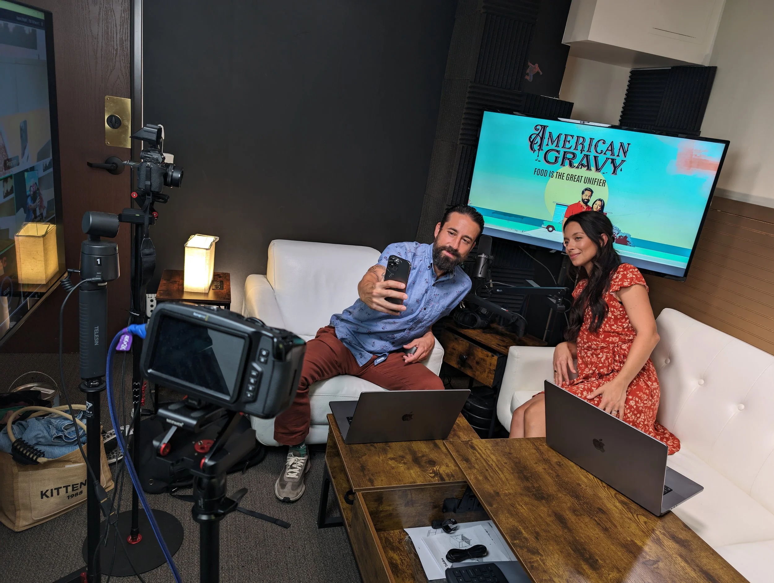 Two people sitting on a white couch in a recording studio, taking a selfie. The man has a beard and wears a blue shirt and brown pants, while the woman has long dark hair and wears a red floral dress. There are laptops, microphones, and lighting equi