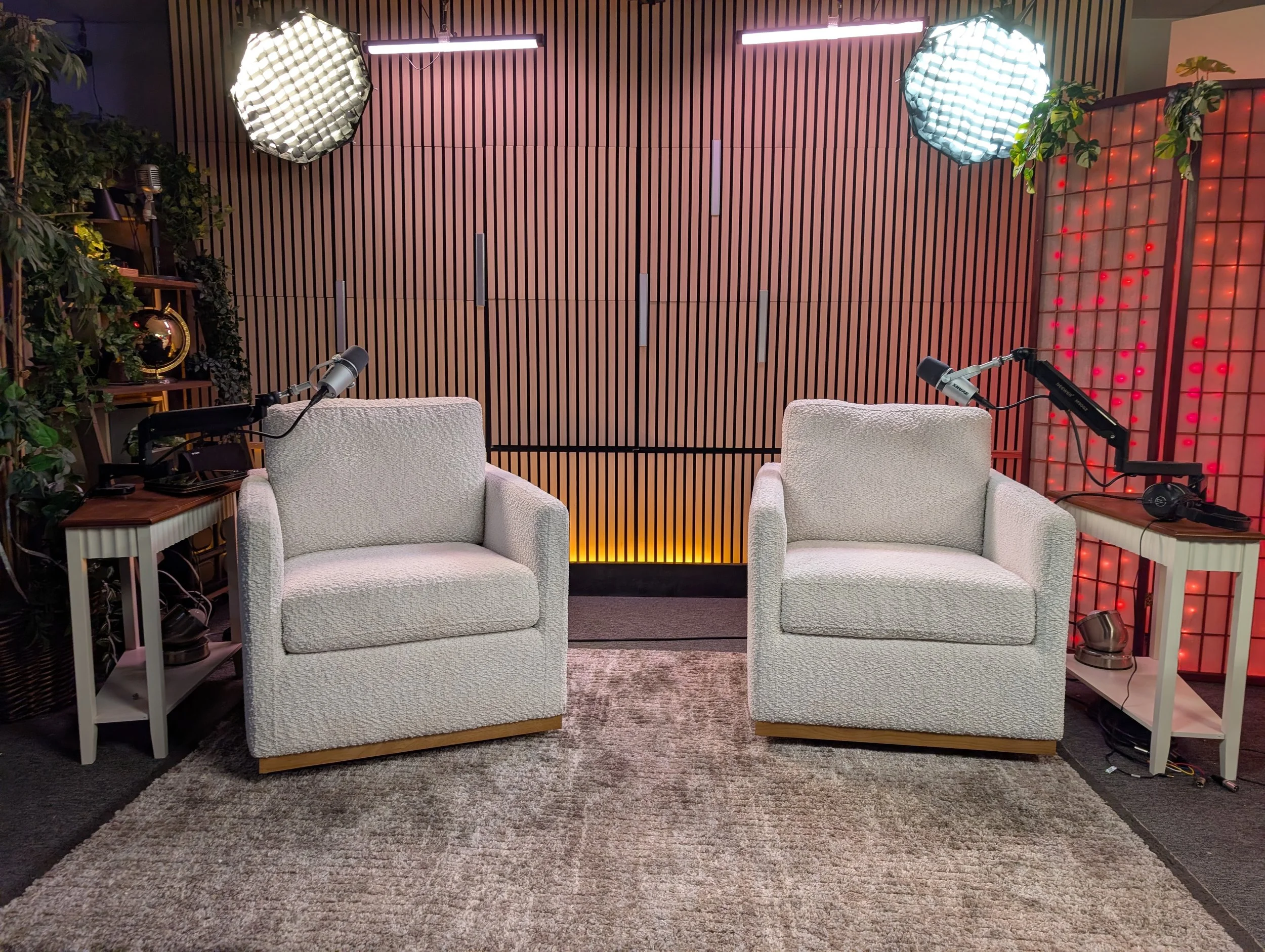 Two white upholstered armchairs with microphones on side tables in a studio setting with wooden wall background, soft lighting, and decorative plants.