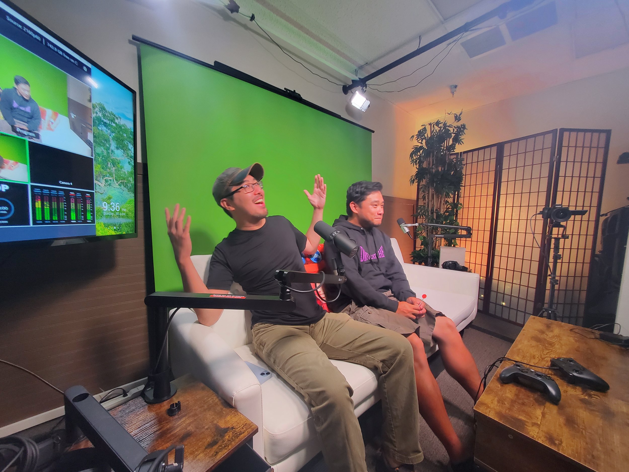 Two men sitting on a white sofa in a recording studio with a green screen behind them, microphones in front of them, gaming controllers on a wooden table, and video equipment around. One man is laughing with his hands raised, the other is sitting qui