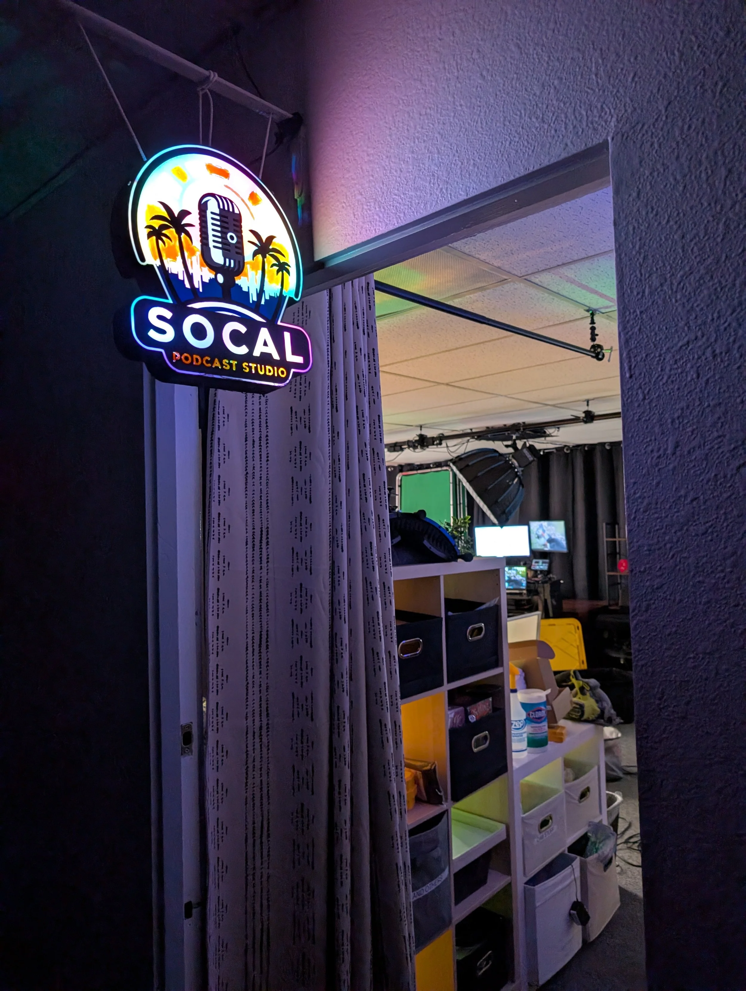 Neon sign that reads 'SOCAL Podcast Studio' with a microphone and palm trees design, inside a recording studio room with equipment and storage bins.
