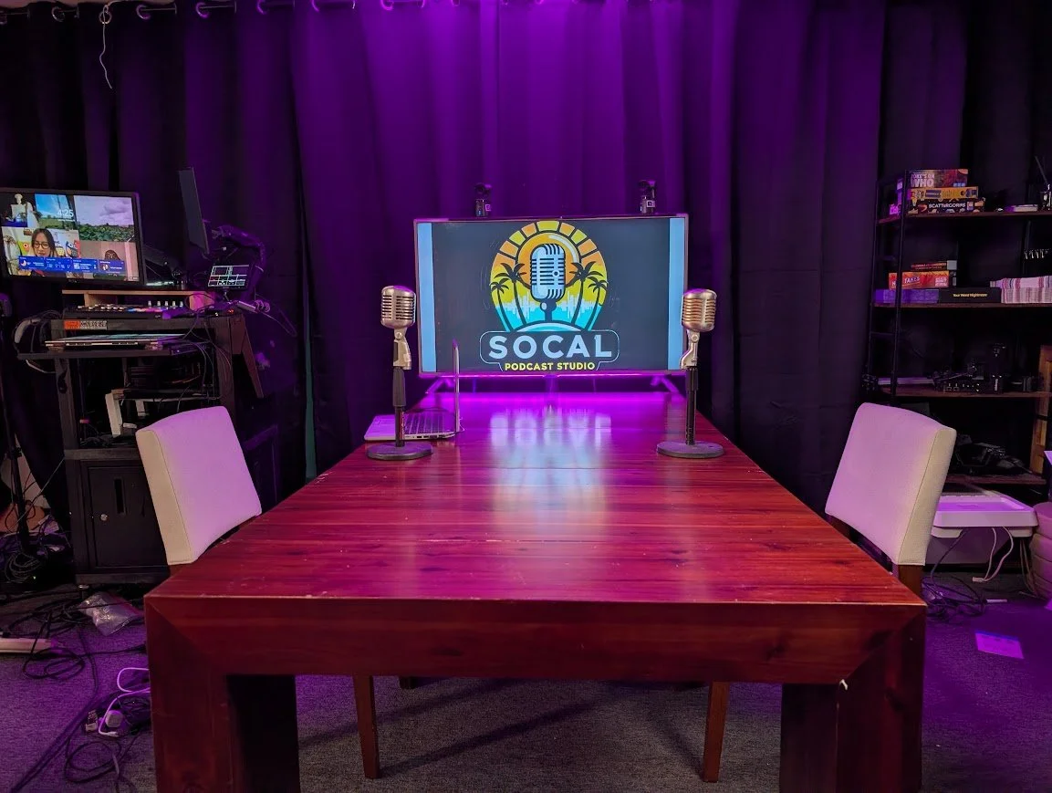 Podcast studio setup with a wooden table, two microphones, a flat-screen with the SoCal Podcast Studio logo, black curtains, and various electronic equipment.