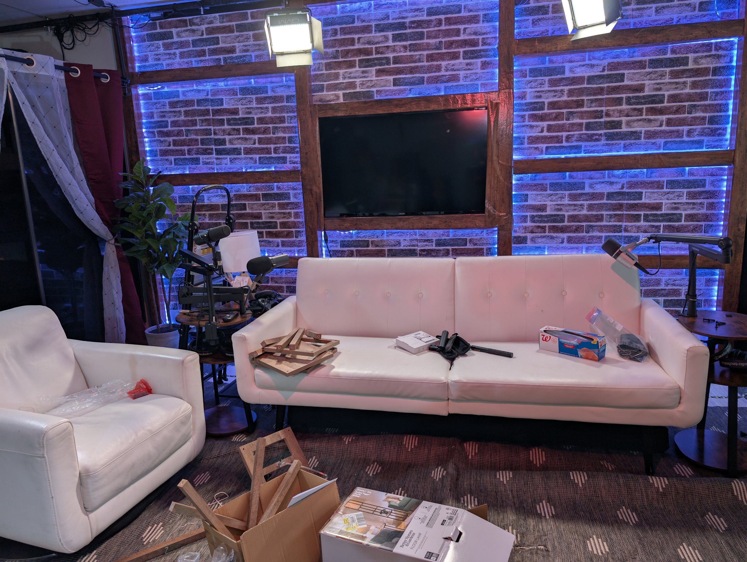 Living room with a white couch, a matching armchair, brick wall with blue LED lighting, a mounted flat-screen TV, and various unpacked items and tools on the furniture and floor.