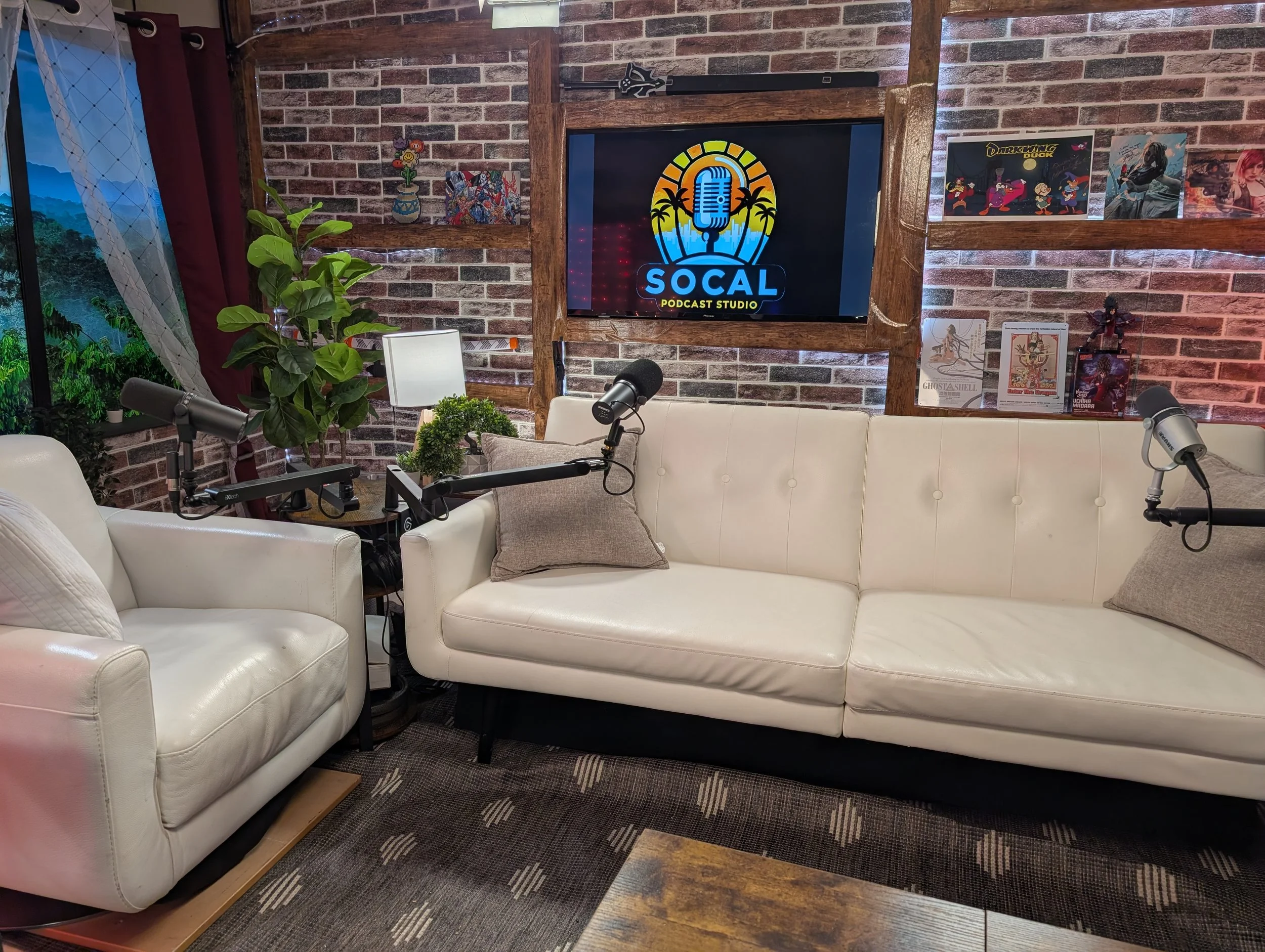 Interior of a podcast studio with a brick wall, a large screen displaying the 'SOCAL Podcast Studio' logo, white couches with pillows, a small table with plants, microphones, and posters on the wall.