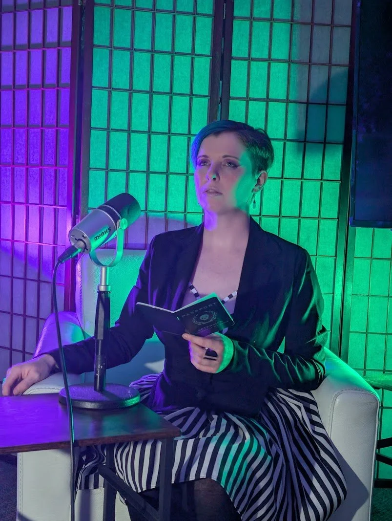 Woman with short hair sitting on a white couch, holding a booklet, with a vintage microphone on a side table, in front of a colorful, illuminated background.