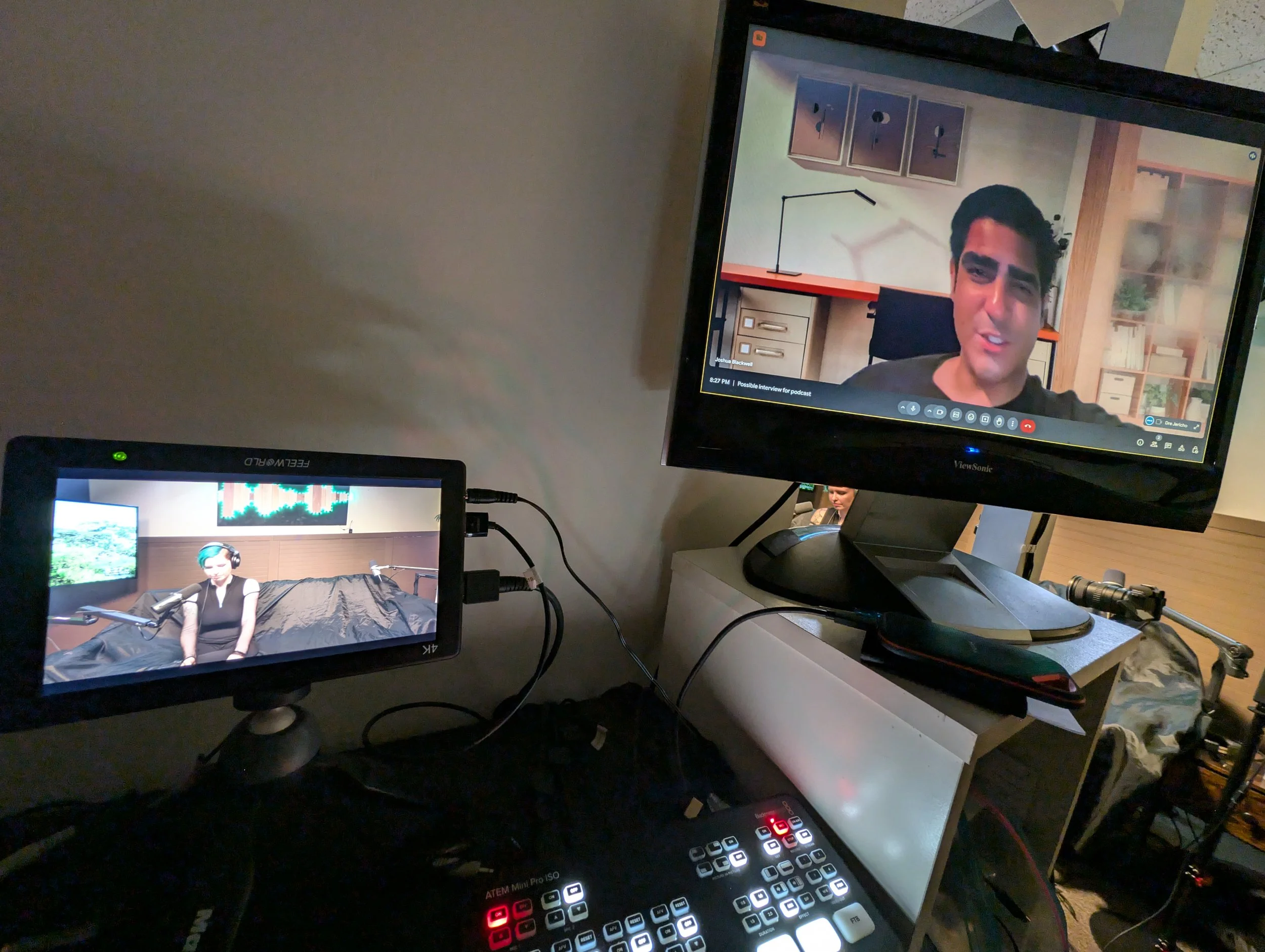 A multi-screen video conferencing setup with an adult woman on one screen wearing headphones and a black shirt, and a man on another screen with dark hair and eyebrows in a home office background. The screens are connected to a control panel with but