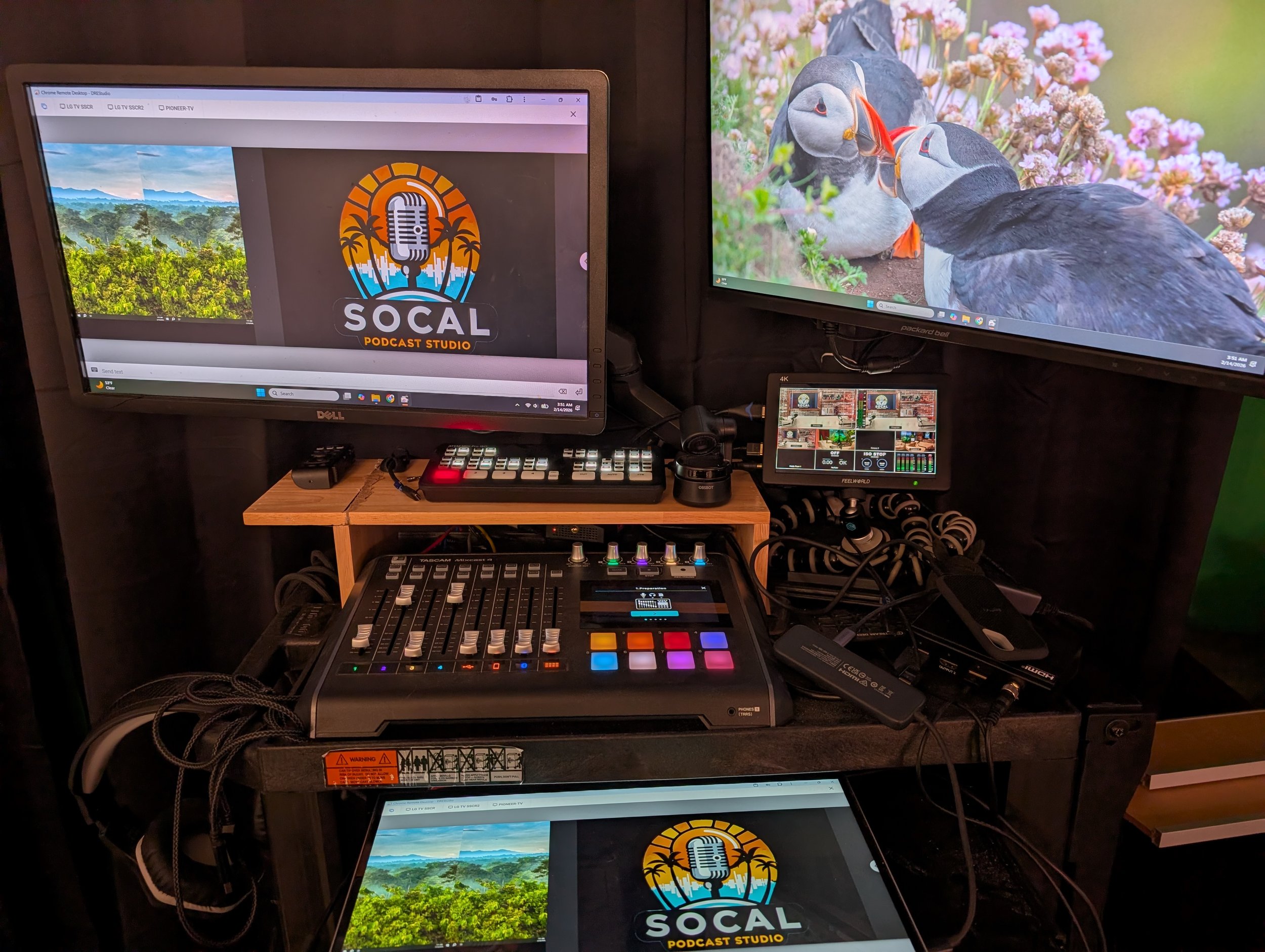 A podcast studio setup with multiple screens, audio equipment, and a logo for SO CAL Podcast Studio on the monitors, including two large screens displaying a landscape and puffin birds, a mixer, and various electronic devices and cables.