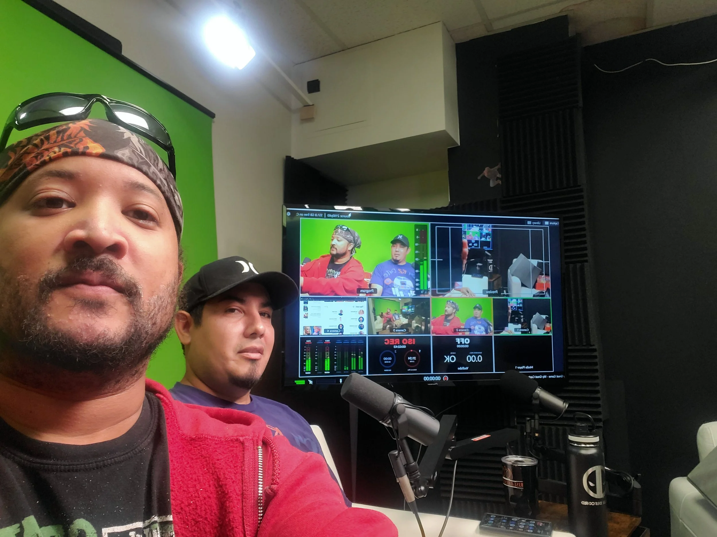 Two men sitting in front of a monitor and microphone in a recording studio, with a green screen background and soundproof walls.