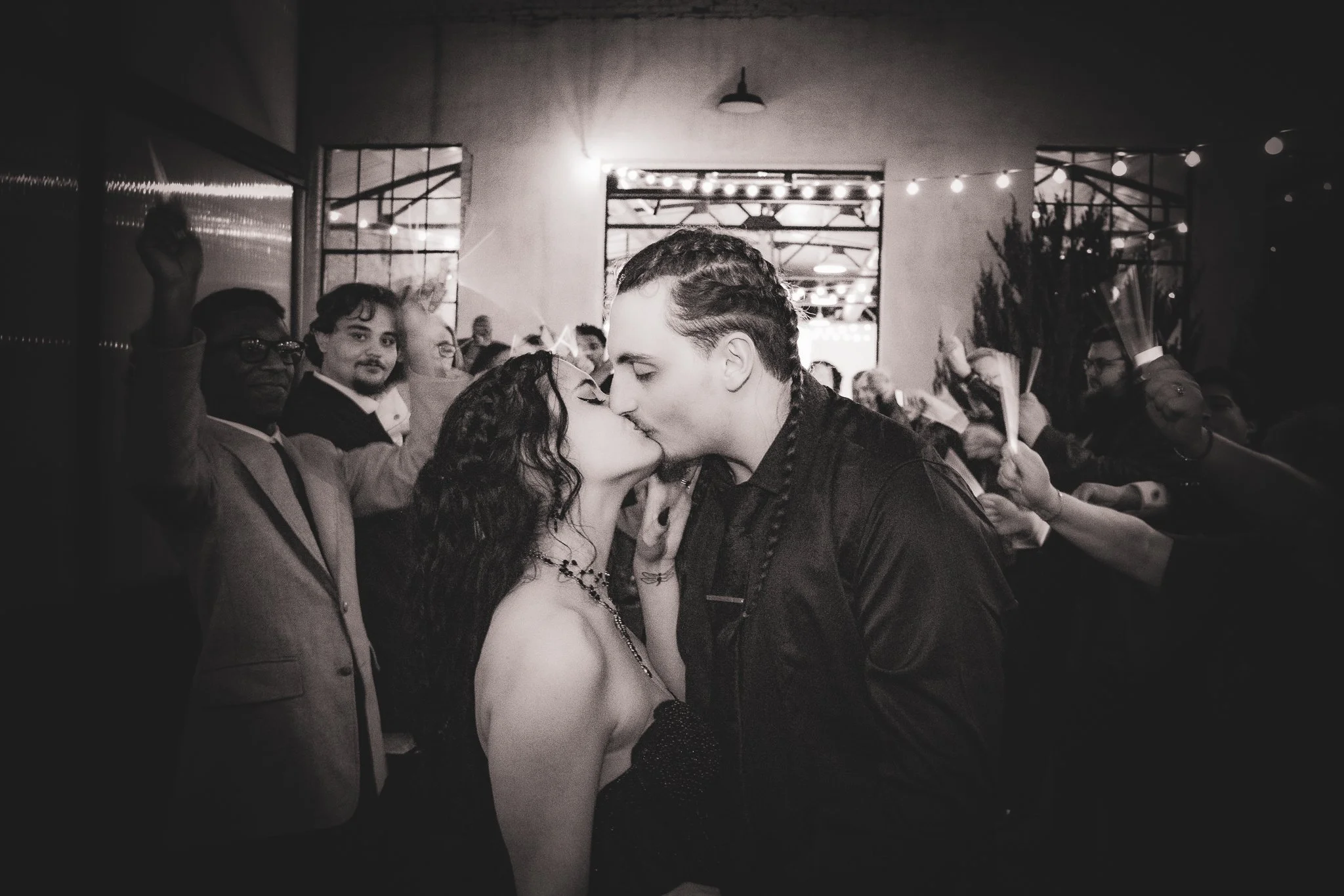 A black and white photo of a couple kissing in the center of a crowd at a celebration or event, with people cheering and holding candles or light sticks in the background.