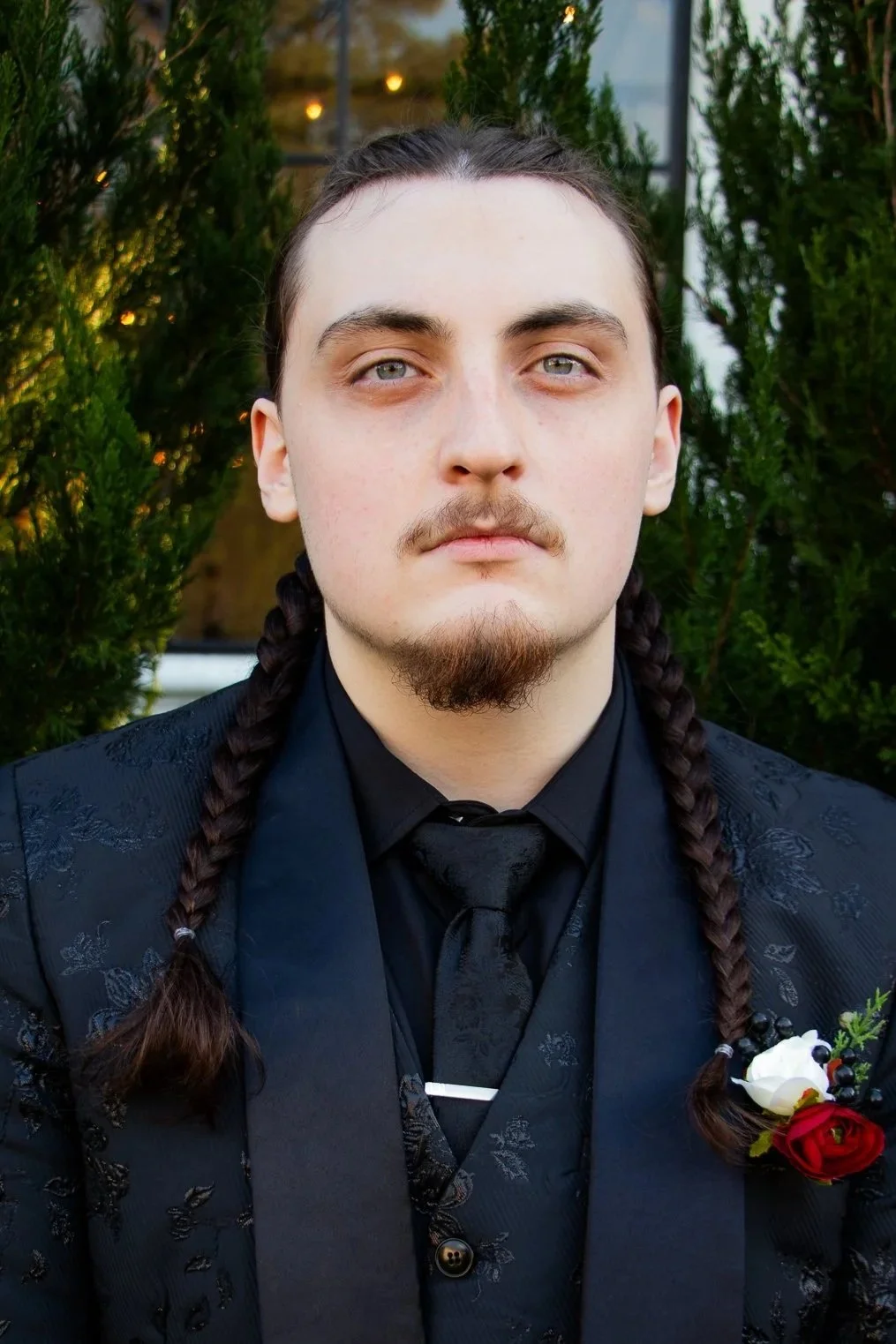 A man with long braided hair, wearing a black suit with subtle floral patterns, a black shirt, and a black tie, standing outdoors against green bushes and trees with blurred lights in the background.