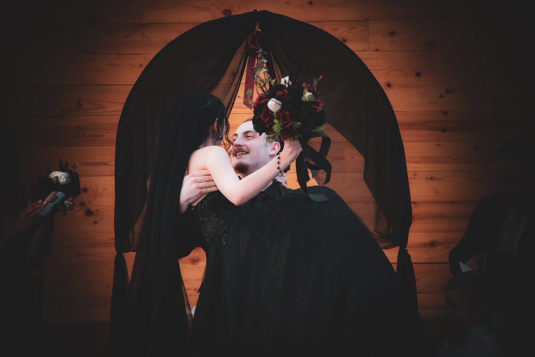A couple dancing at their wedding, with the groom holding the bride in his arms. The bride is holding a bouquet of dark red, white, and green flowers. They are standing under a black sheer fabric canopy, with a wooden wall in the background. Guests a