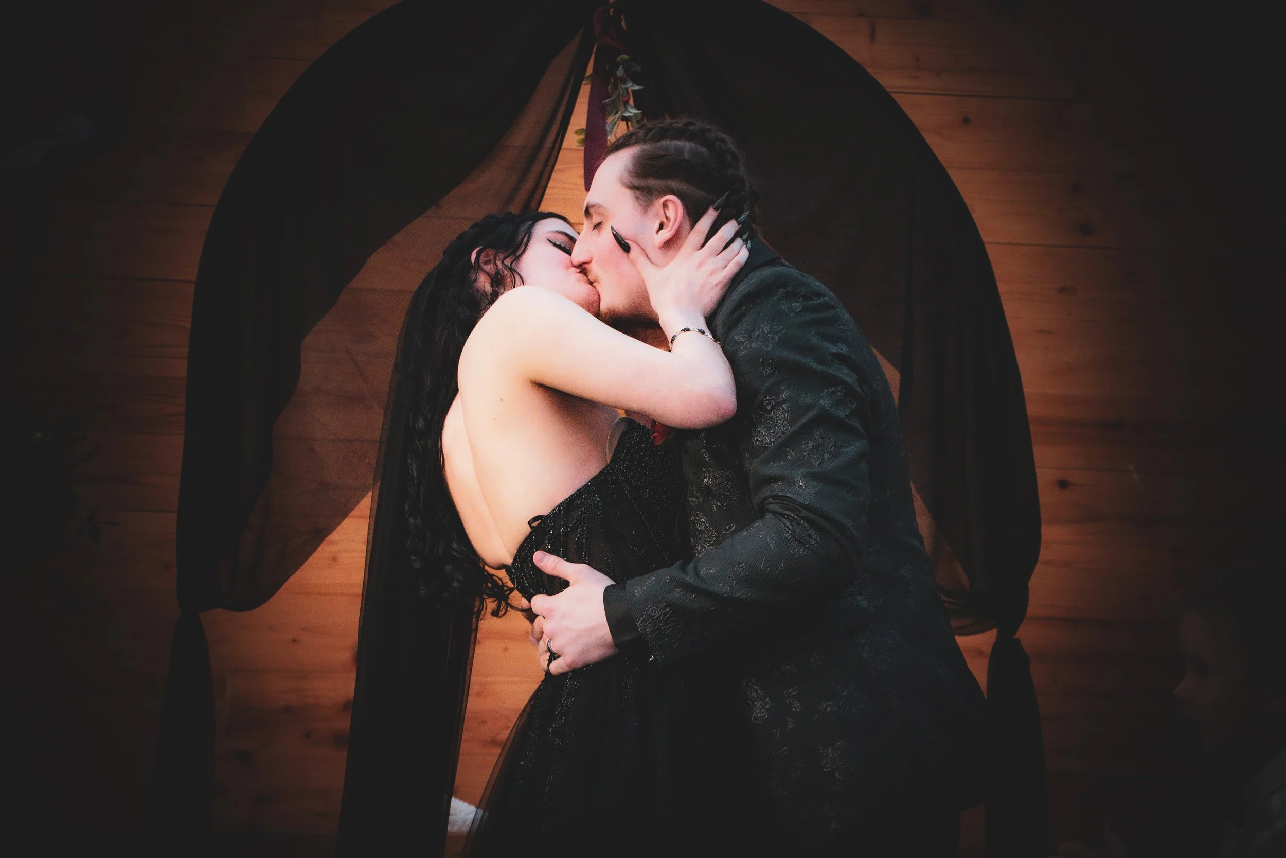 A young couple sharing a passionate kiss, with the woman wearing a black dress and the man in a dark outfit, against a wooden wall background.