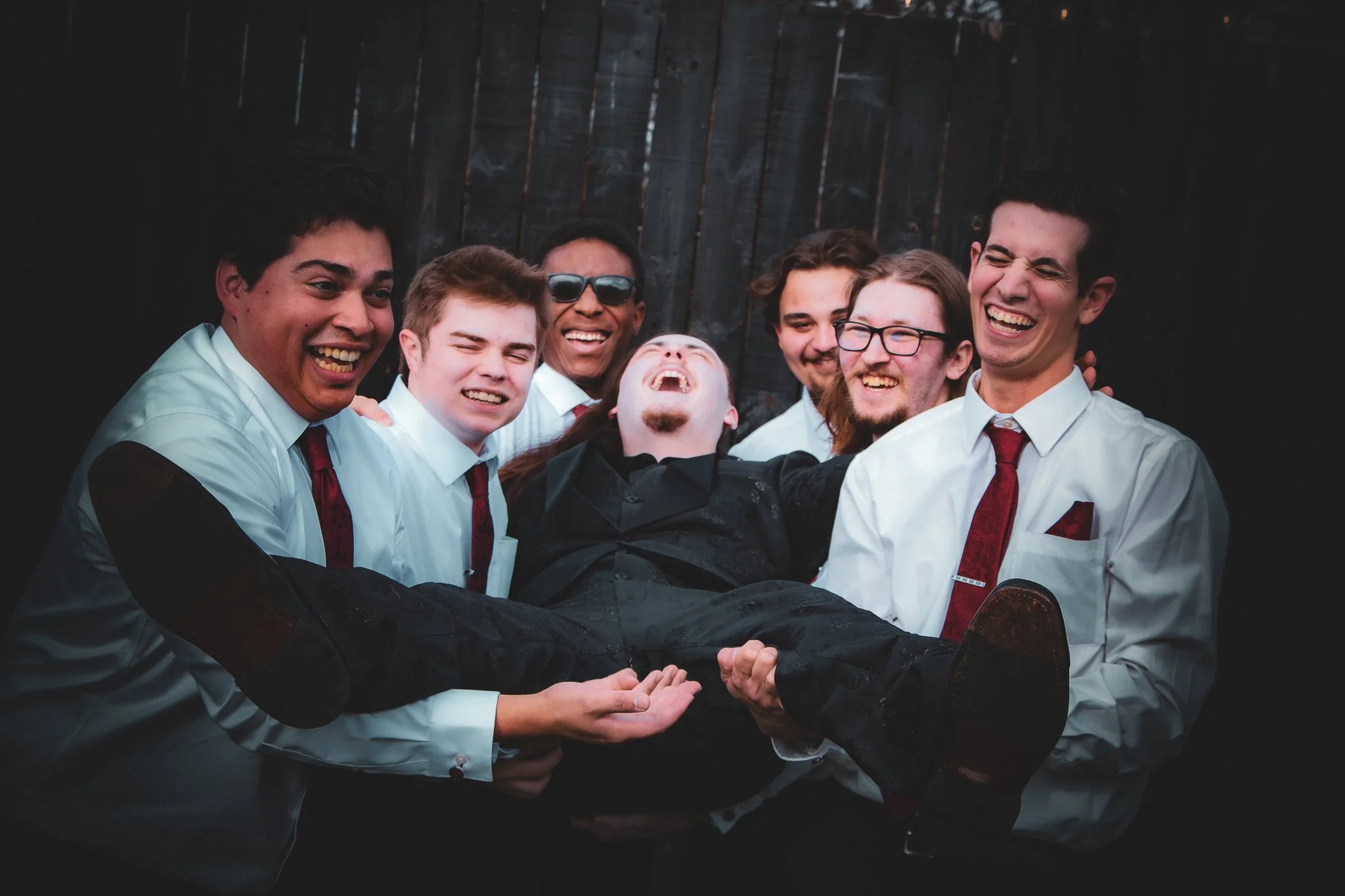 Group of six men lifting a man in a black shirt, all smiling and laughing, in front of a dark wooden fence.