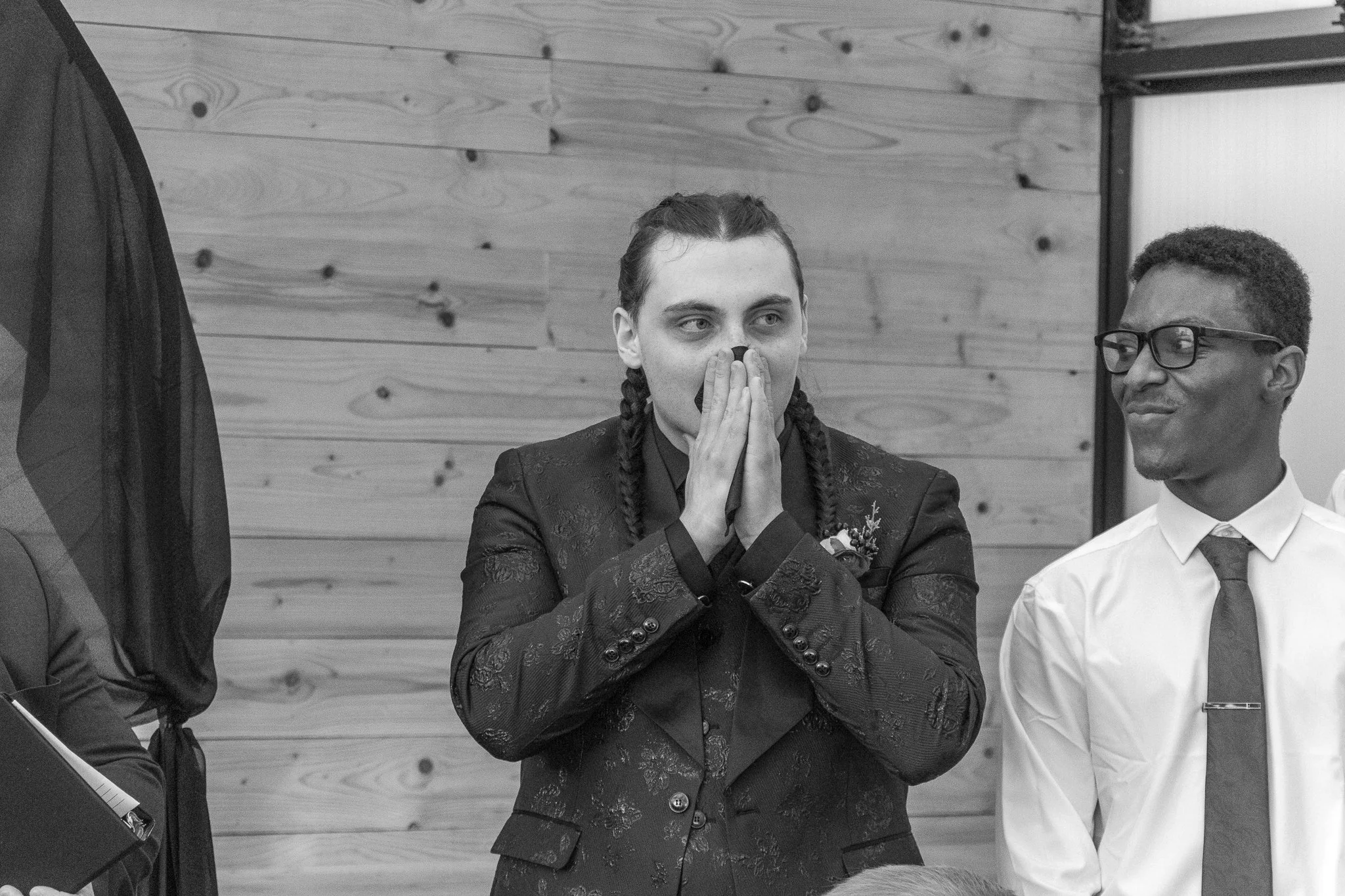 A woman with braided hair, wearing a patterned blazer, has her hands over her face as if emotional. She is indoors with a wooden wall in the background. To her right, a man wearing glasses, a white shirt, and a tie is looking at her with a smile.