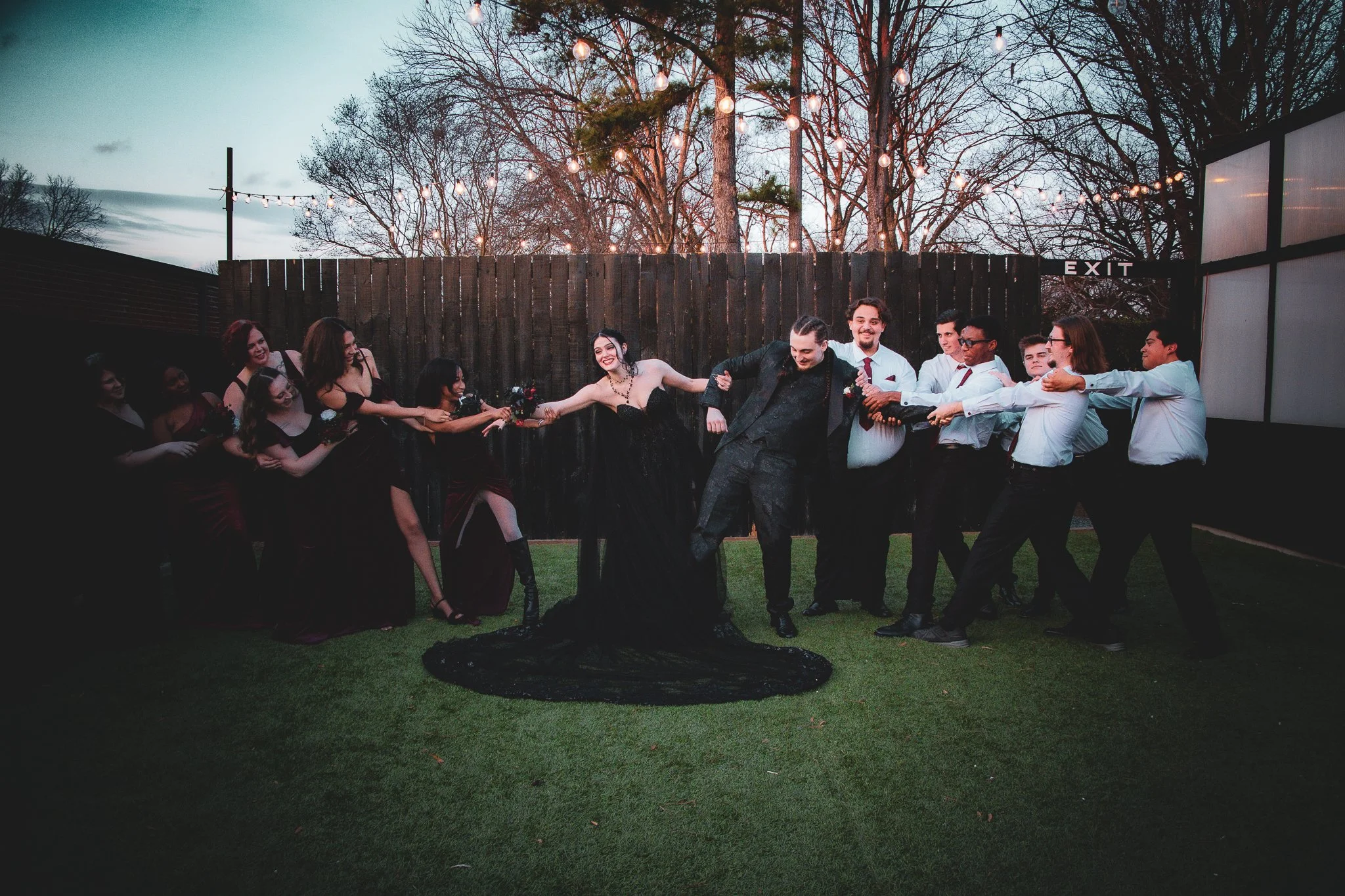 Group of people at a wedding or celebration playing tug-of-war game with a bride and groom in the center outdoors during evening.
