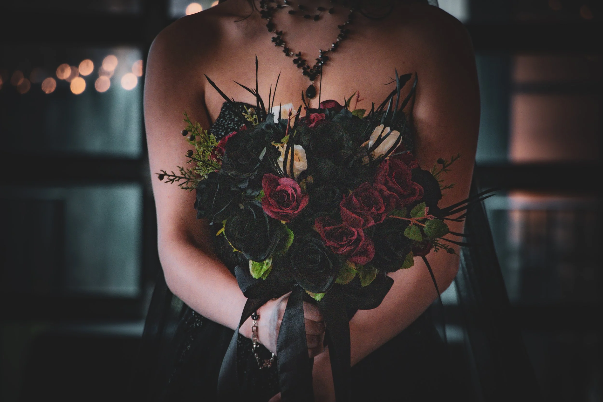 A woman in a strapless black dress holding a bouquet of dark red and black roses with greenery, in a dimly lit setting.