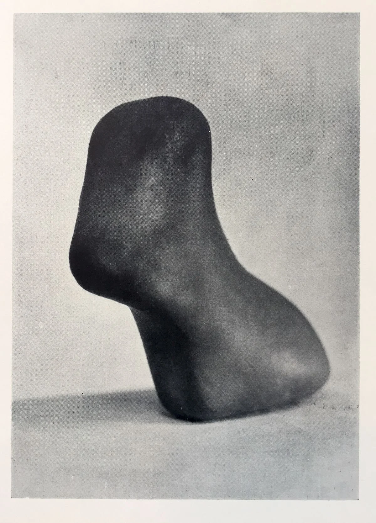 Black and white photo of a smooth, abstract sculpture resembling a rounded figure with a curved extension, placed on a flat surface.
