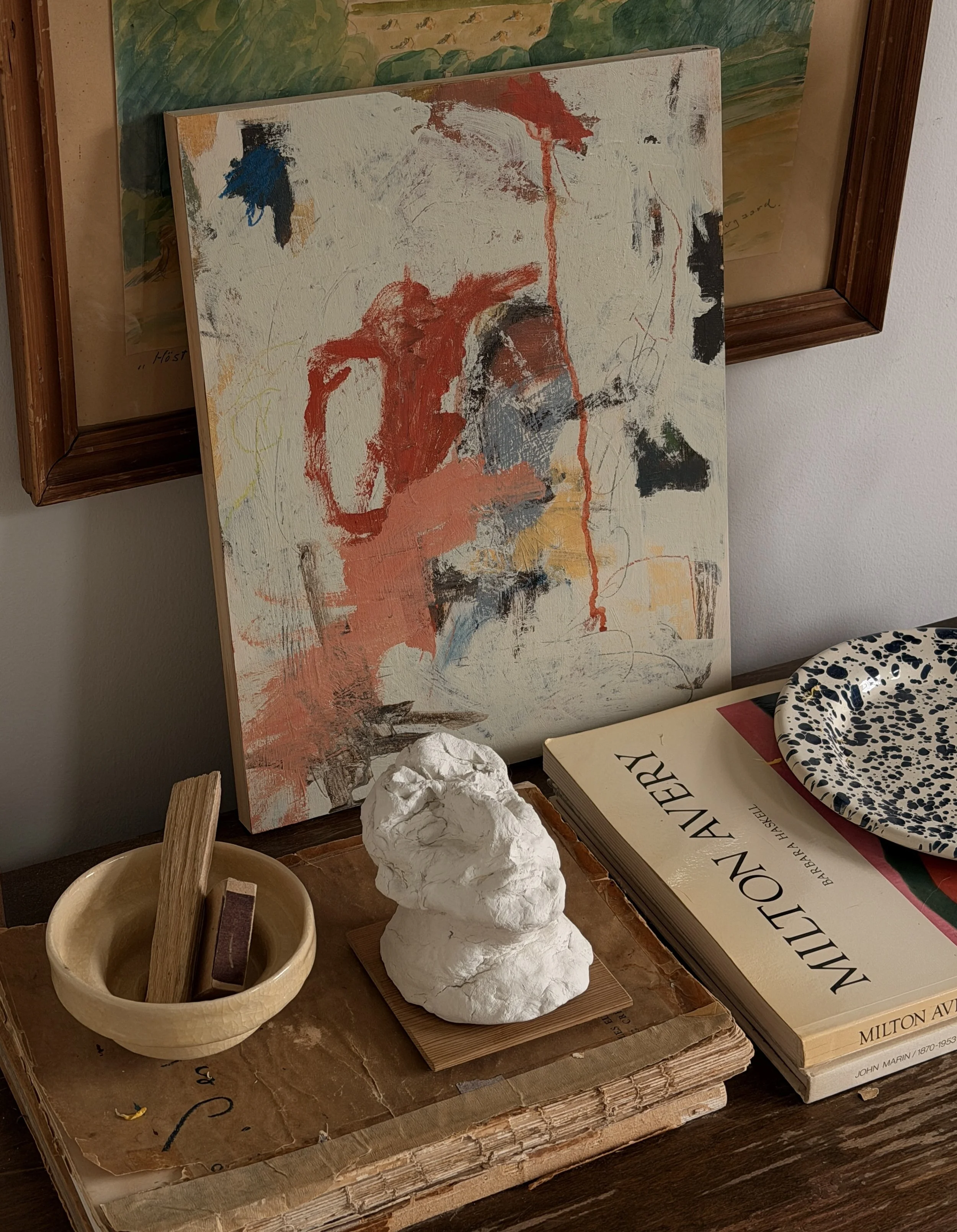 Interior of an art gallery or studio displaying a large abstract painting with splashes of red, black, and blue on a cream background, leaning against the wall. On the table below, there is a white, irregularly shaped sculpture, a wooden bowl with a plaster pestle, and a large art book titled 'Milton Avery'.