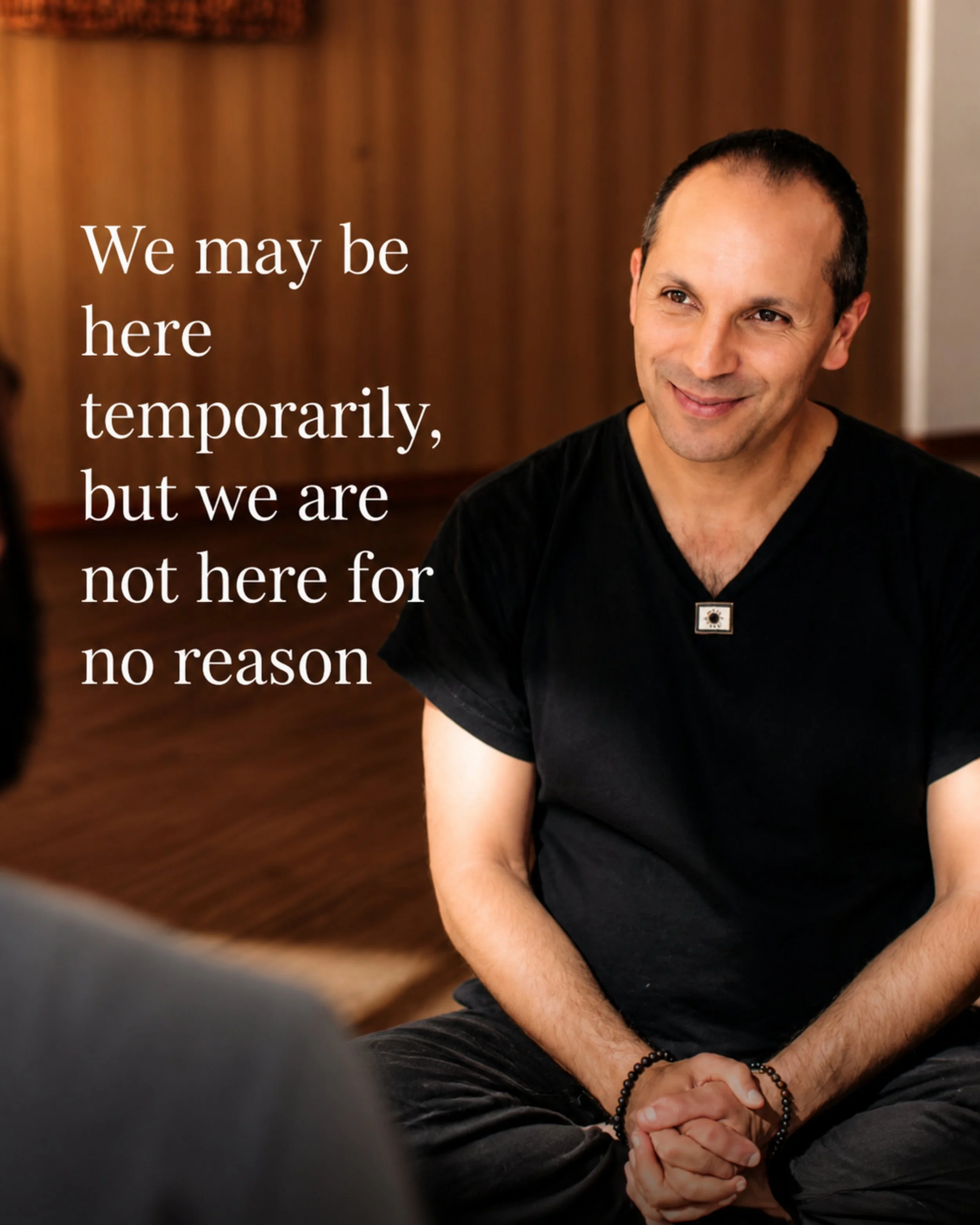Close-up of a smiling man sitting cross-legged with his hands clasped in his lap, wearing a black shirt and beaded bracelet, in a room with wooden paneling.