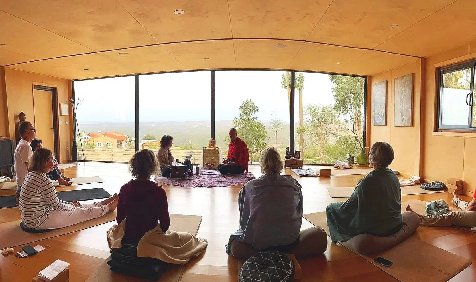 People participating in a meditation or yoga session in a spacious room with large floor-to-ceiling windows overlooking a scenic landscape with trees and houses. The participants are seated on cushions or mats, facing a person at the front leading the session.